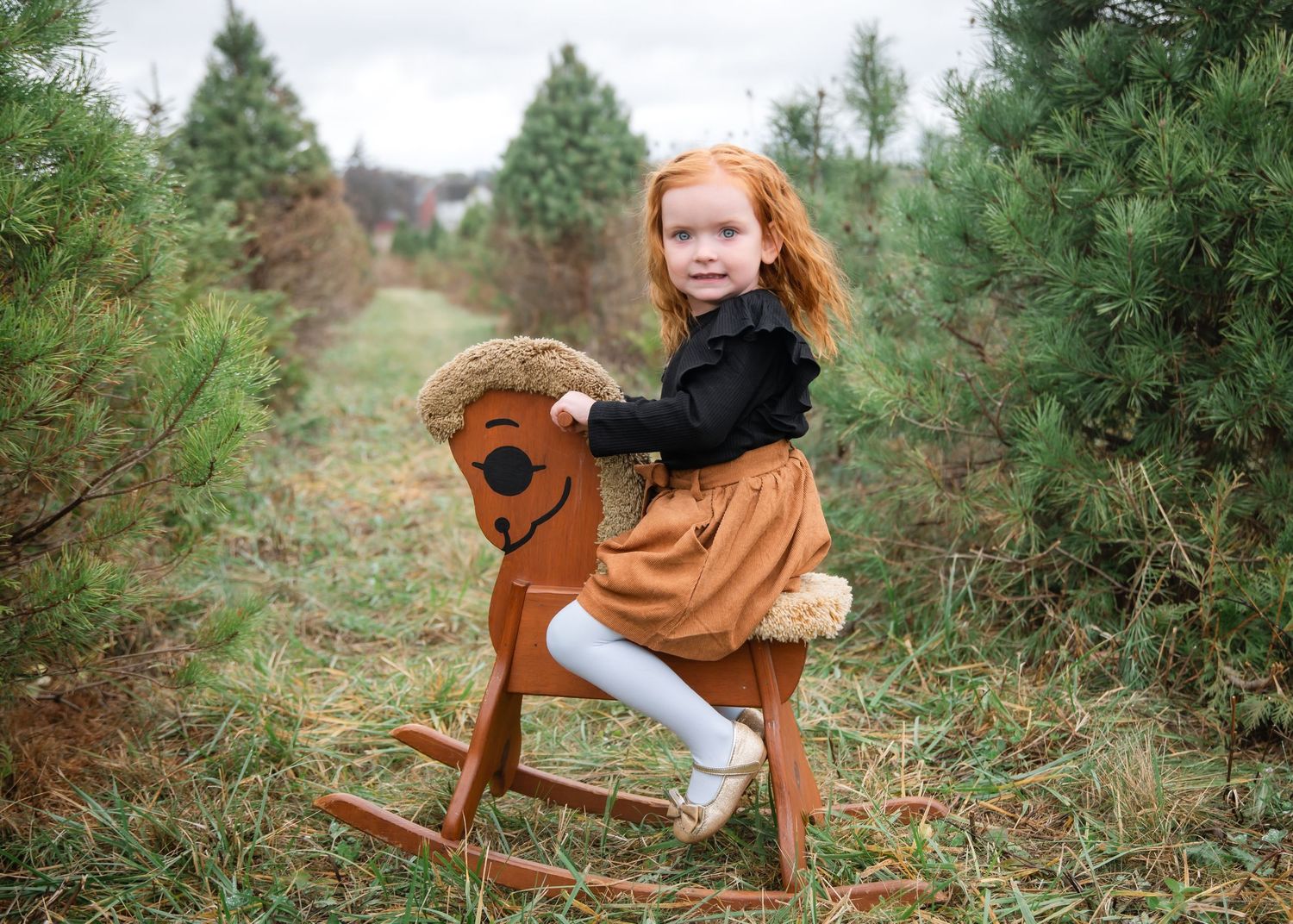 A wooden rocking horse sits among evergreen trees in a rustic outdoor setting during an autumn photoshoot.
