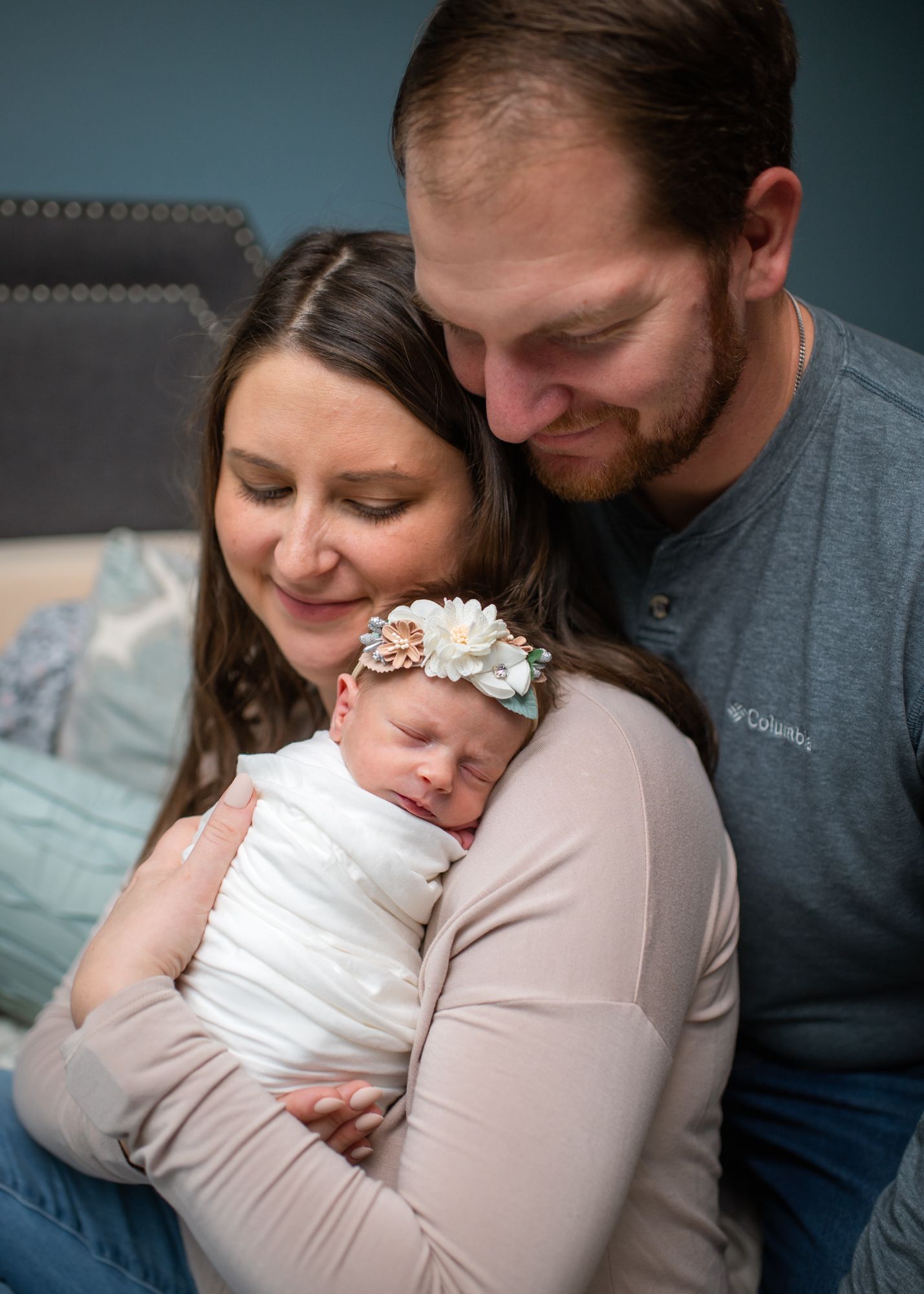 New parents snuggle with sleeping newborn on grey couch during lifestyle photoshoot.