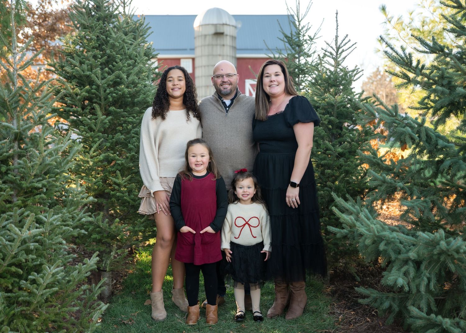 Family of five posing among Christmas trees at a farm, with a red barn in the background on a festive holiday outing.