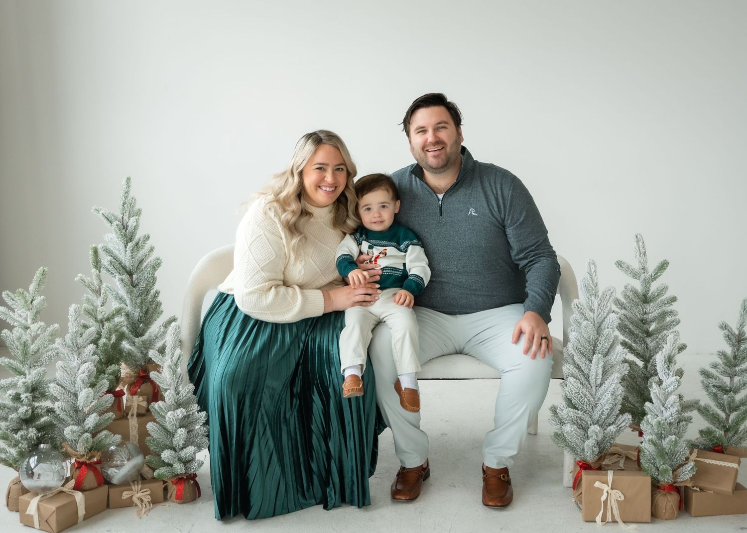 Family of three posing for a Christmas portrait with snowy trees and gift boxes in a white studio.