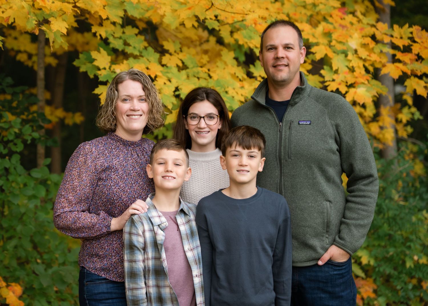 Family of five smiling outdoors in autumn, surrounded by vibrant yellow and orange foliage.