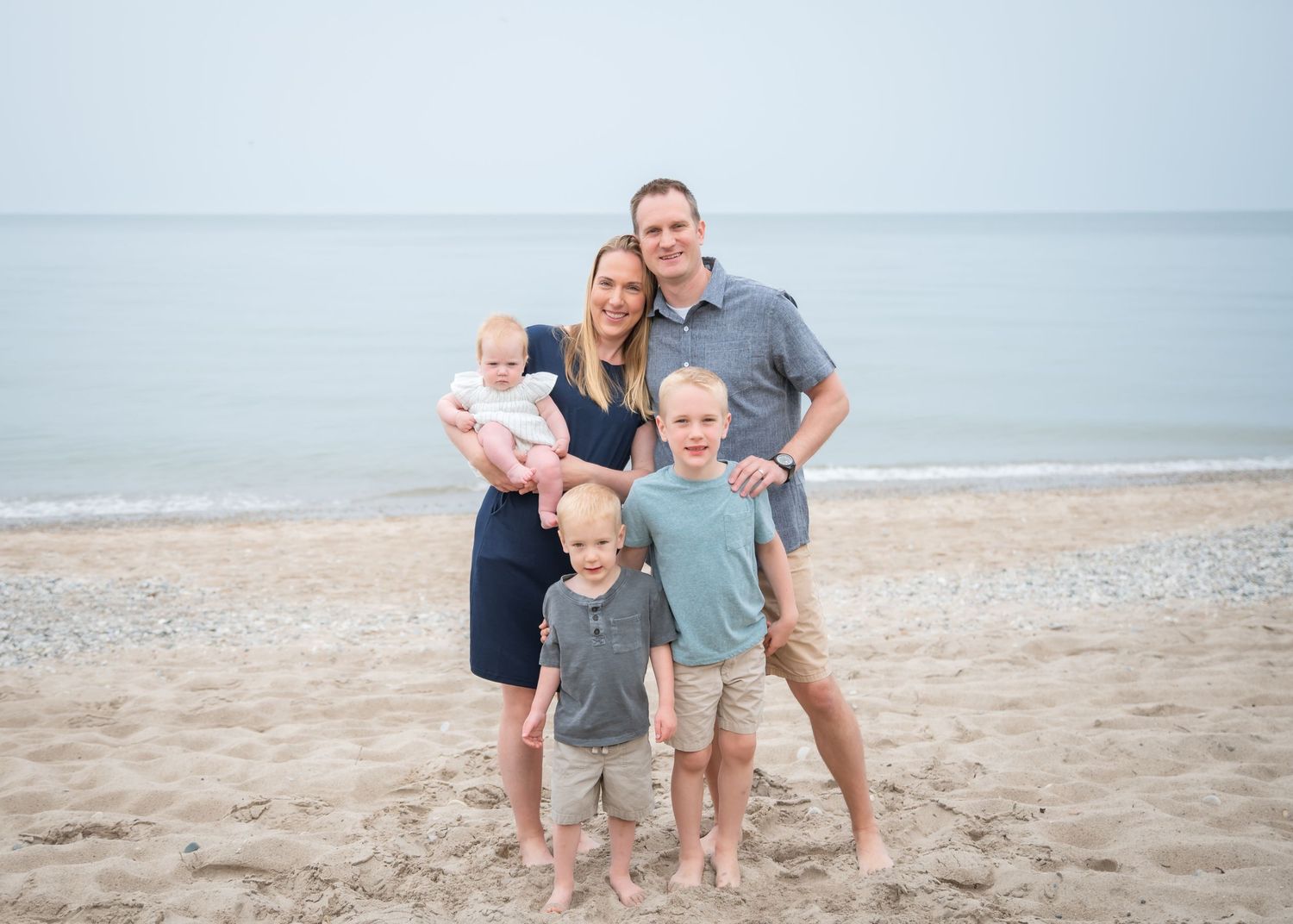 Family of five posing on sandy beach, parents holding baby with two young boys in front, calm ocean backdrop.