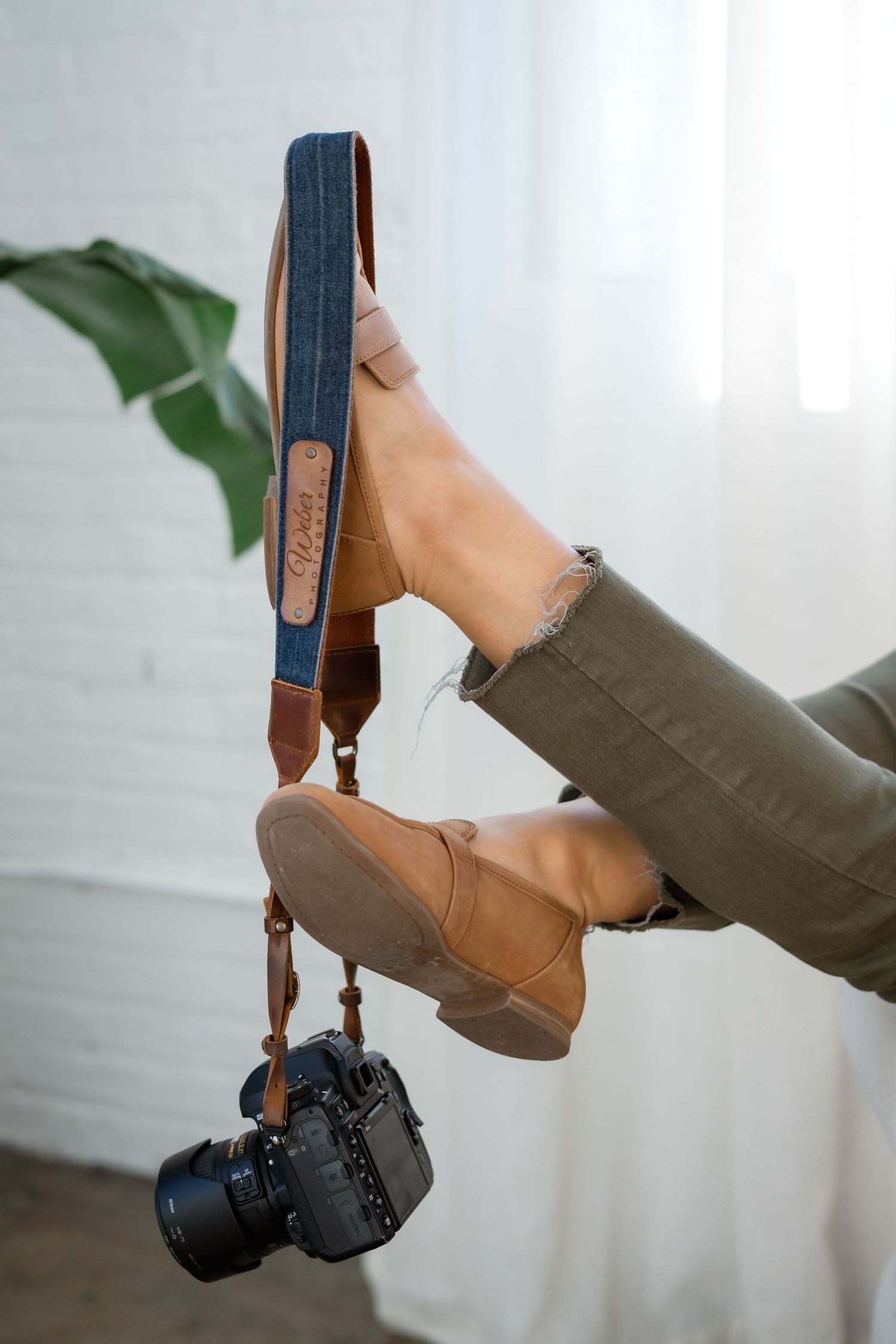 A leather camera strap attached to a DSLR camera hangs from someone's boot in a lifestyle product photo.