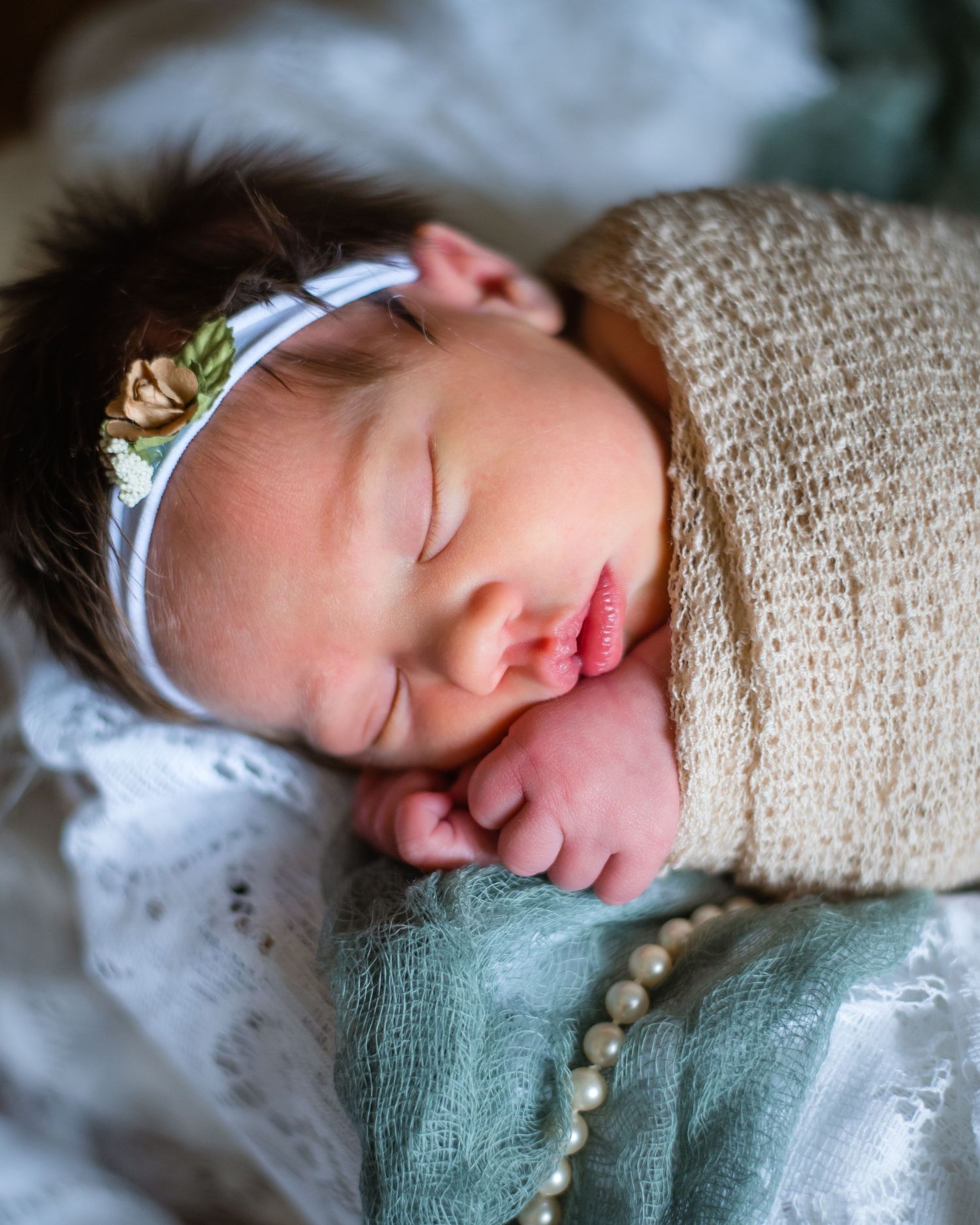 A sleeping newborn with dark hair wears a floral headband against soft textured fabrics.
