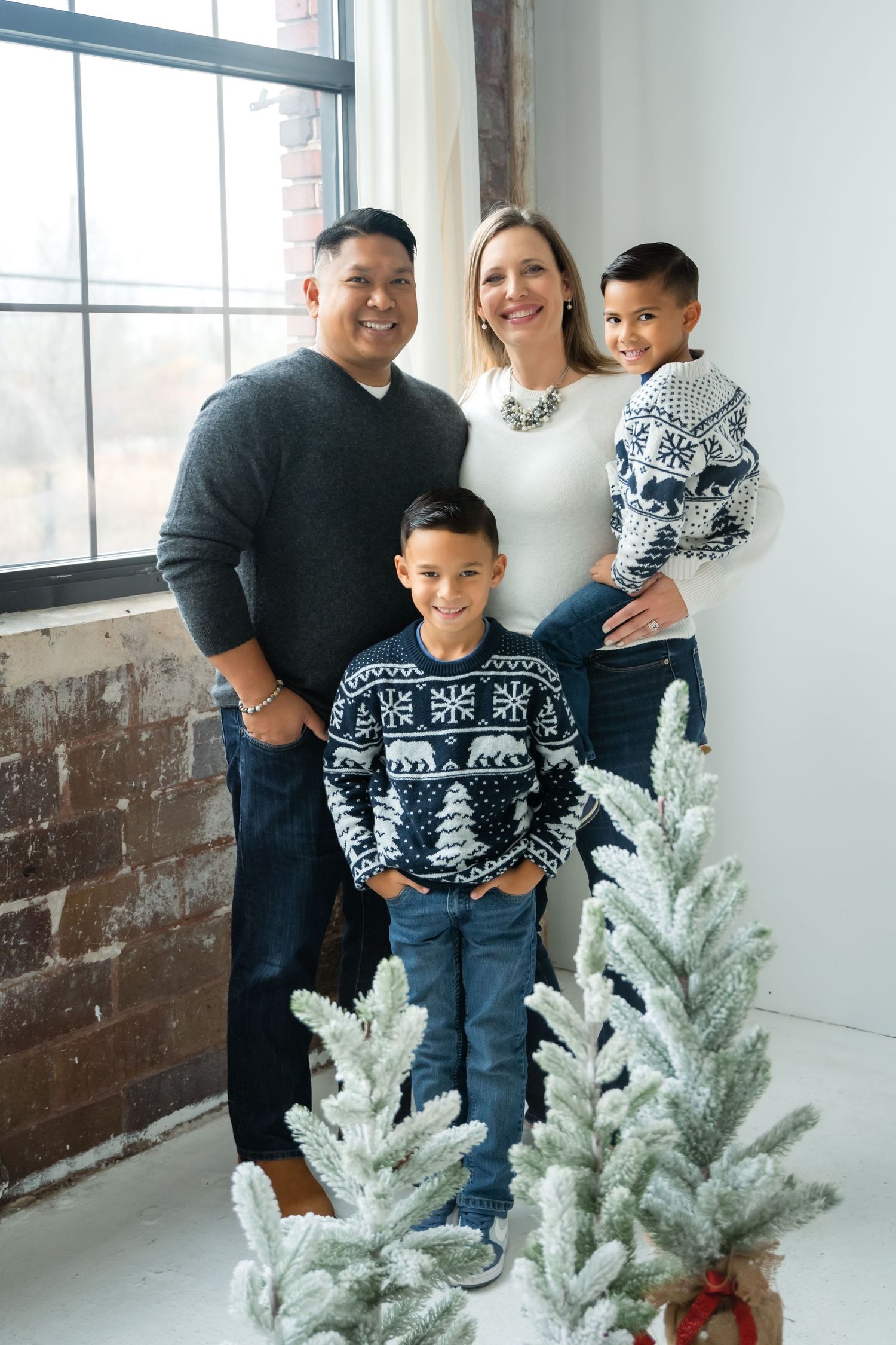 Family of four in holiday sweaters posing with snowy Christmas trees in a bright studio setting.