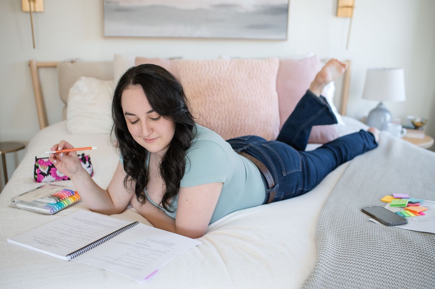Woman lying on bed with open notebook, colorful sticky notes, and pen, planning or studying in cozy bedroom.