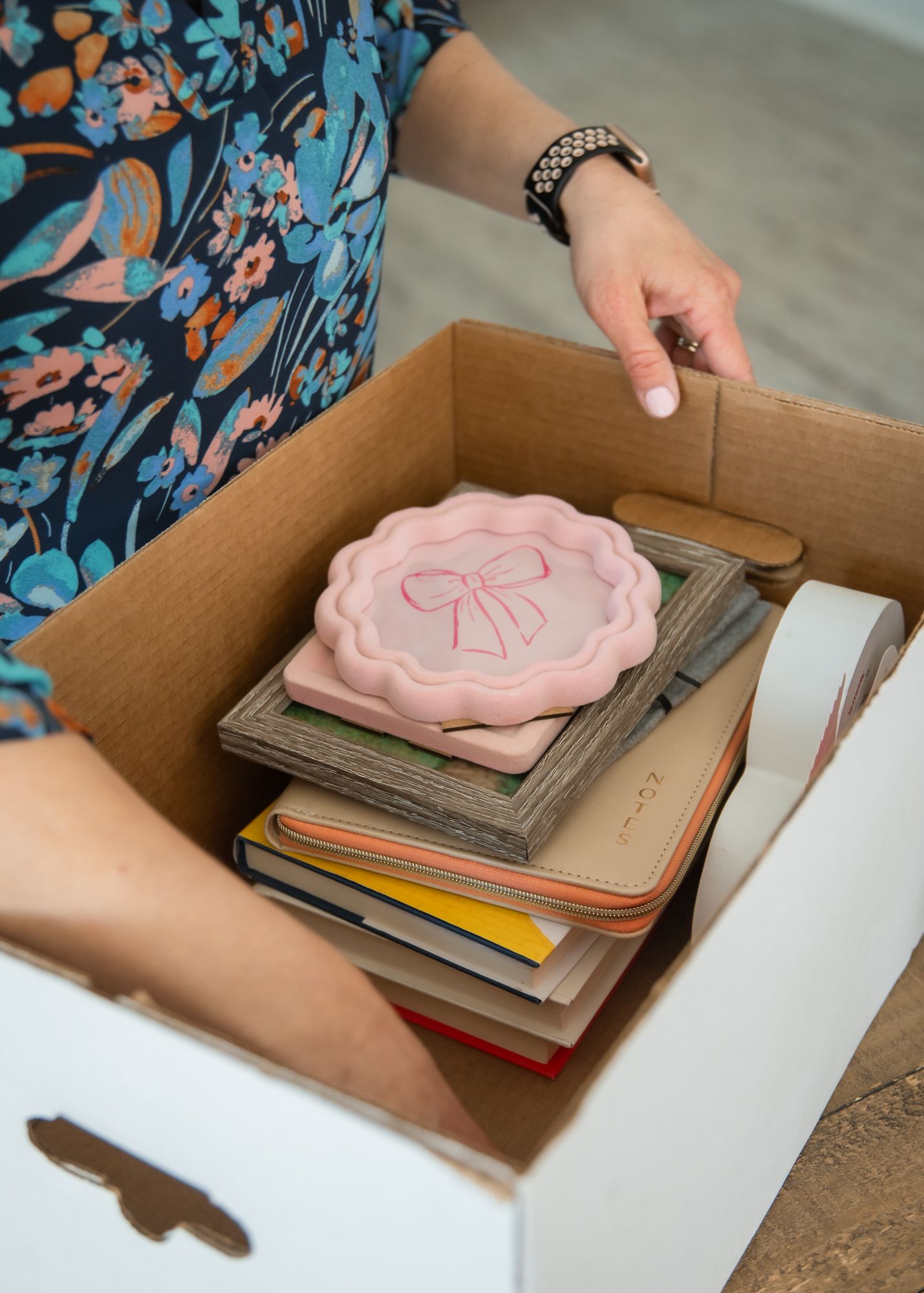 Person placing a pink bow decorative box on top of stacked books and journals inside a cardboard moving box.
