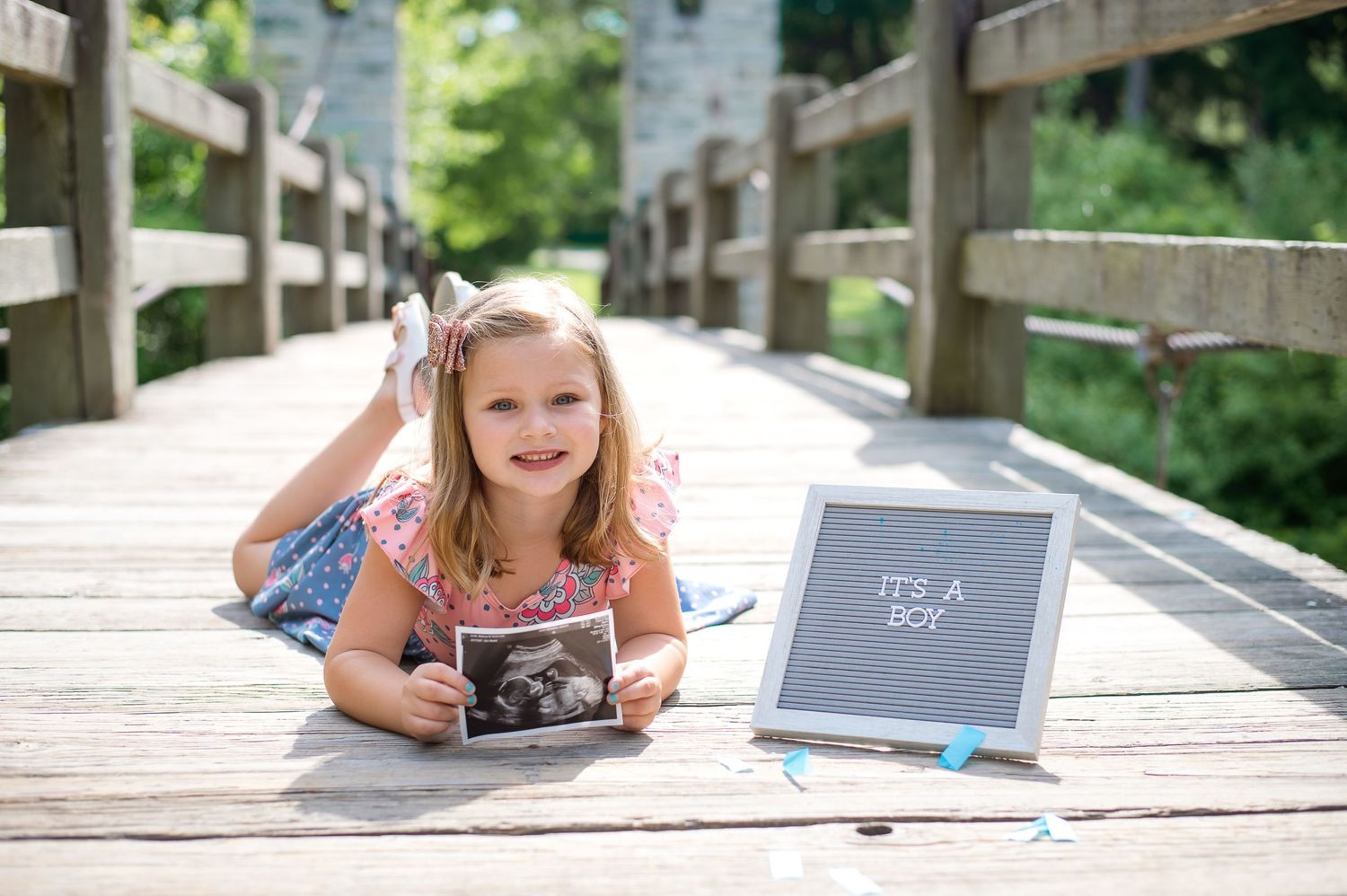 Pregnancy announcement photo with letterboard and ultrasound photo on wooden bridge outdoors.