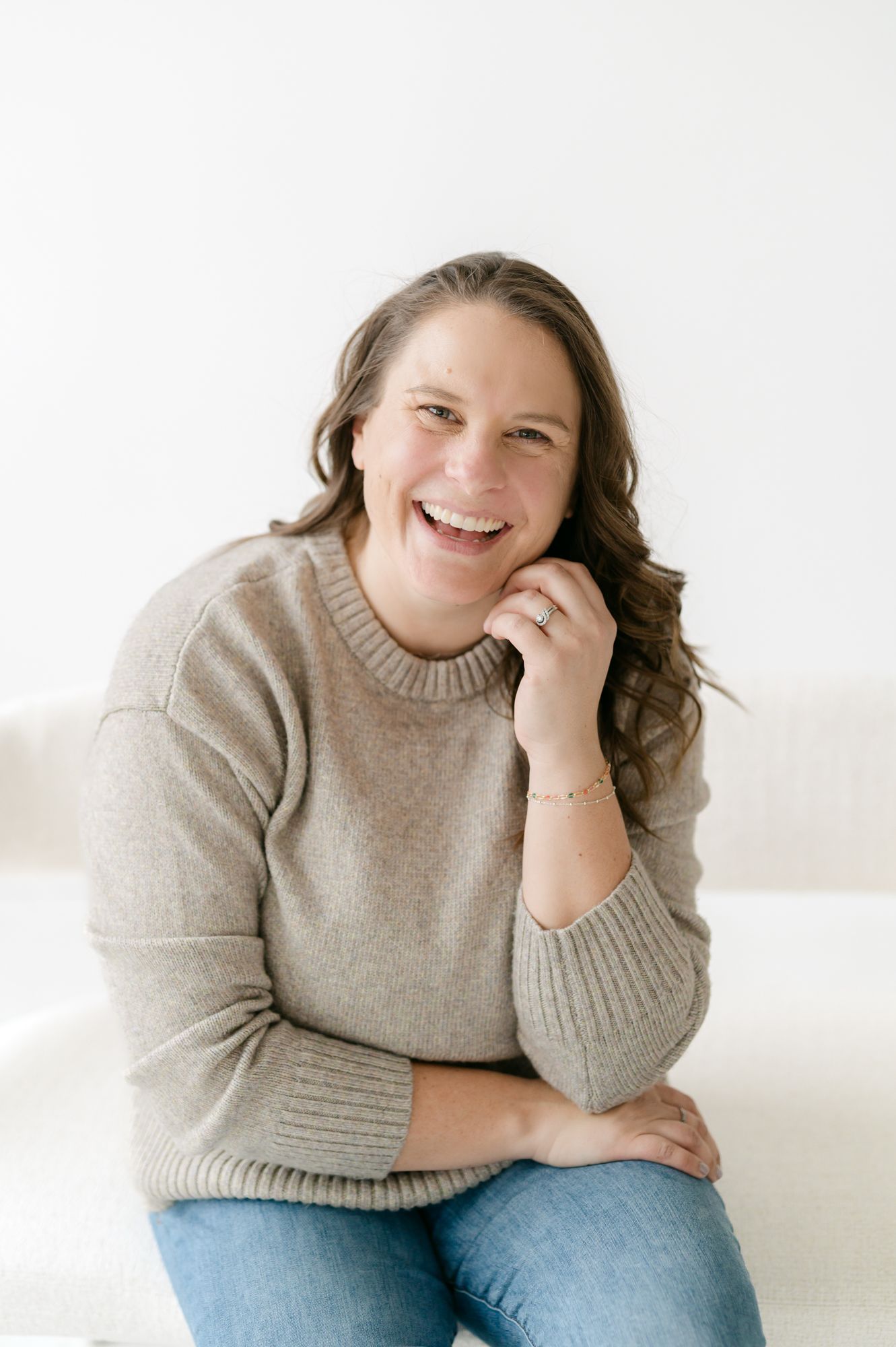 A person wearing a beige sweater and jeans sits casually while laughing and smiling in a bright white studio setting.