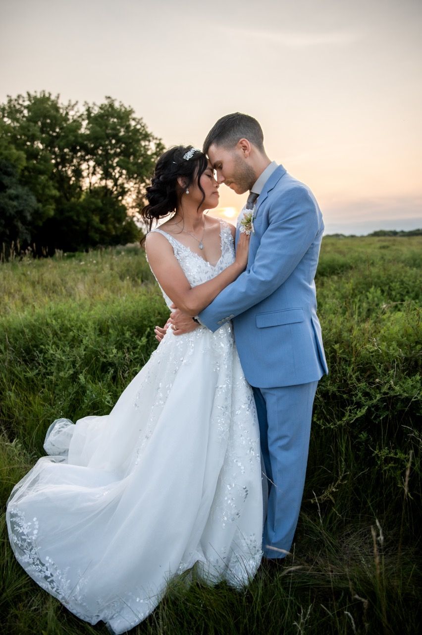 Romantic sunset wedding photo of couple in light blue suit and white lace dress against natural landscape.