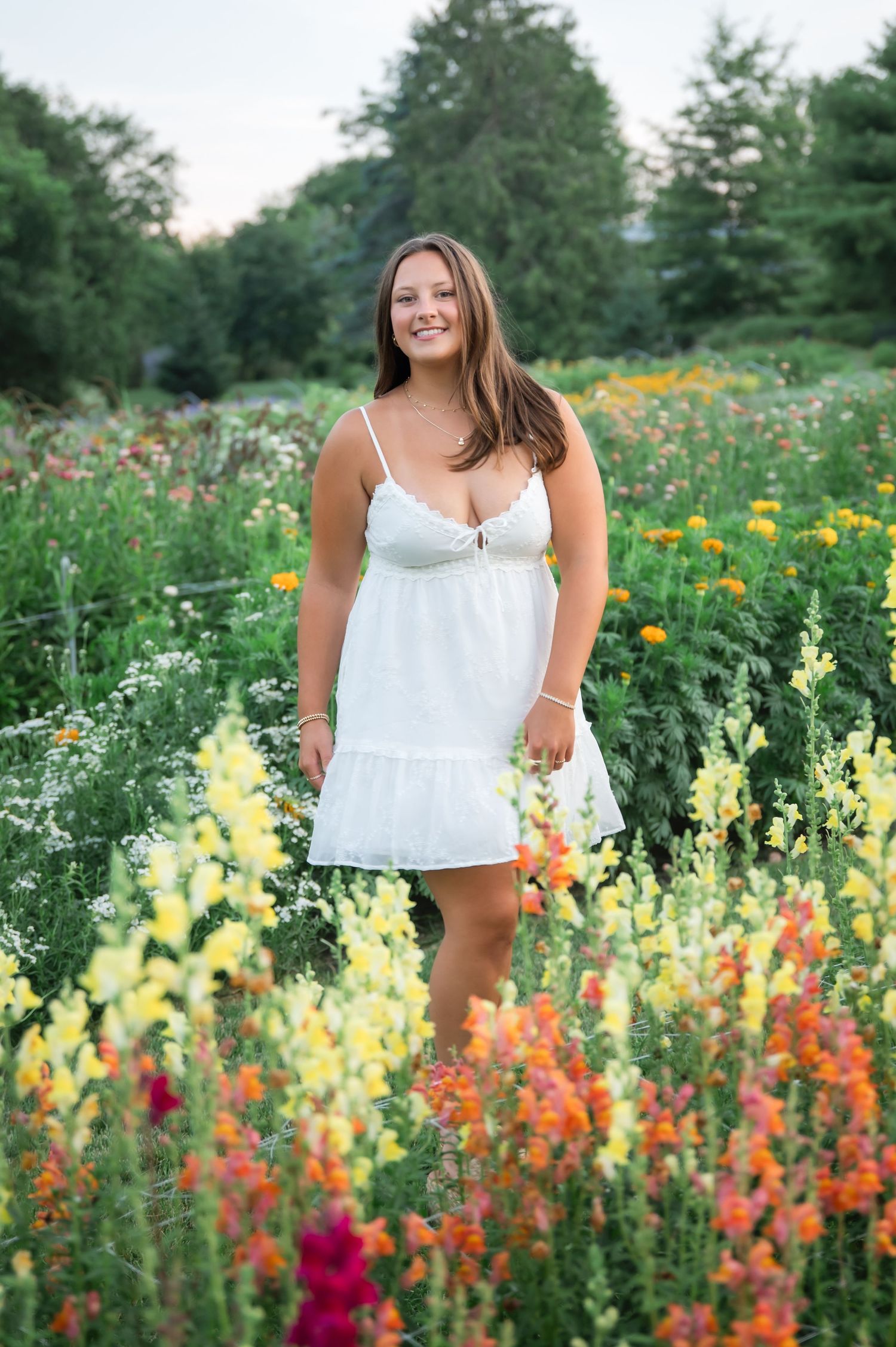 Young woman in white sundress smiling among colorful wildflowers in a lush garden at dusk.