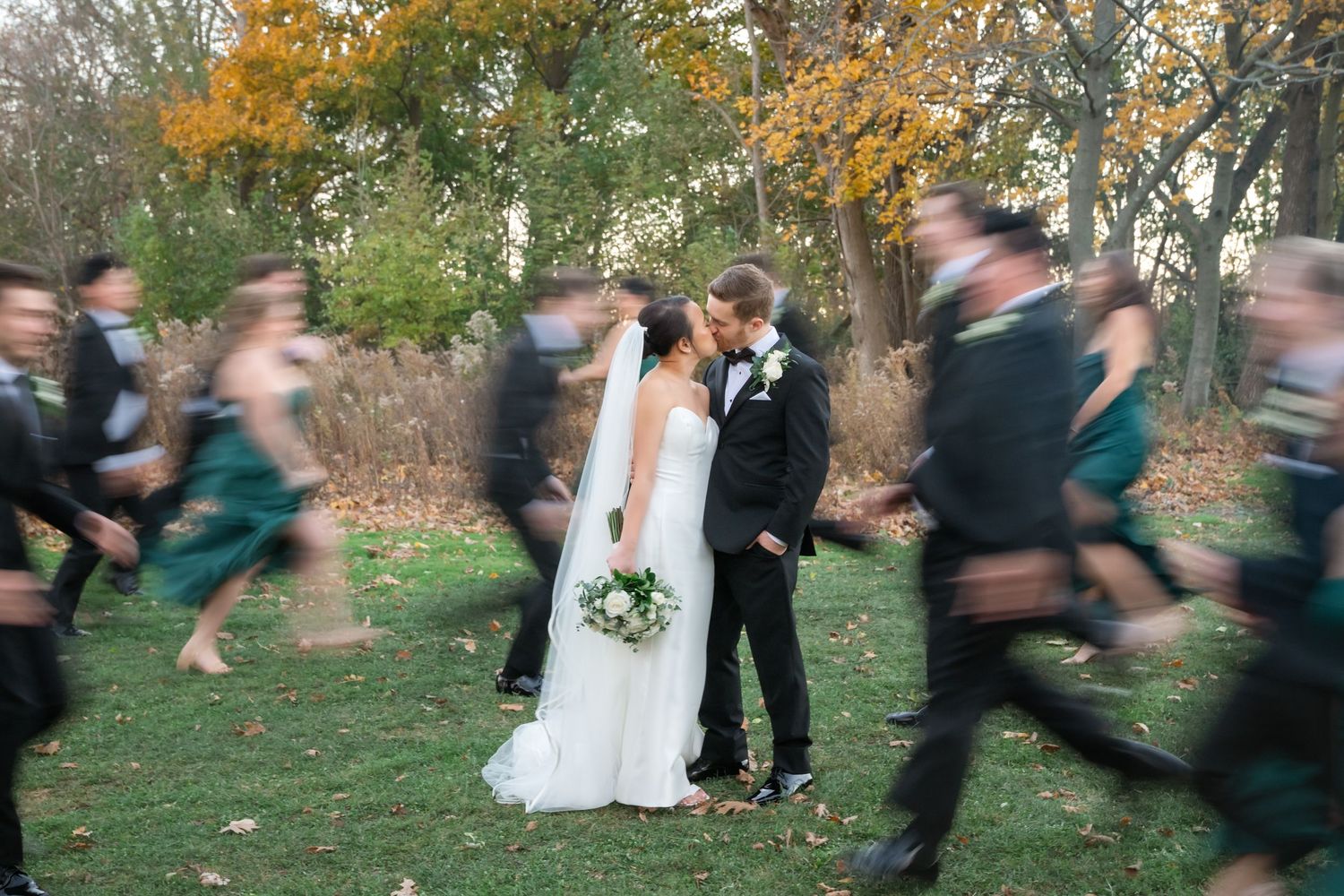 Wedding party members run and blur into motion around a bride and groom standing still in an autumn garden setting.