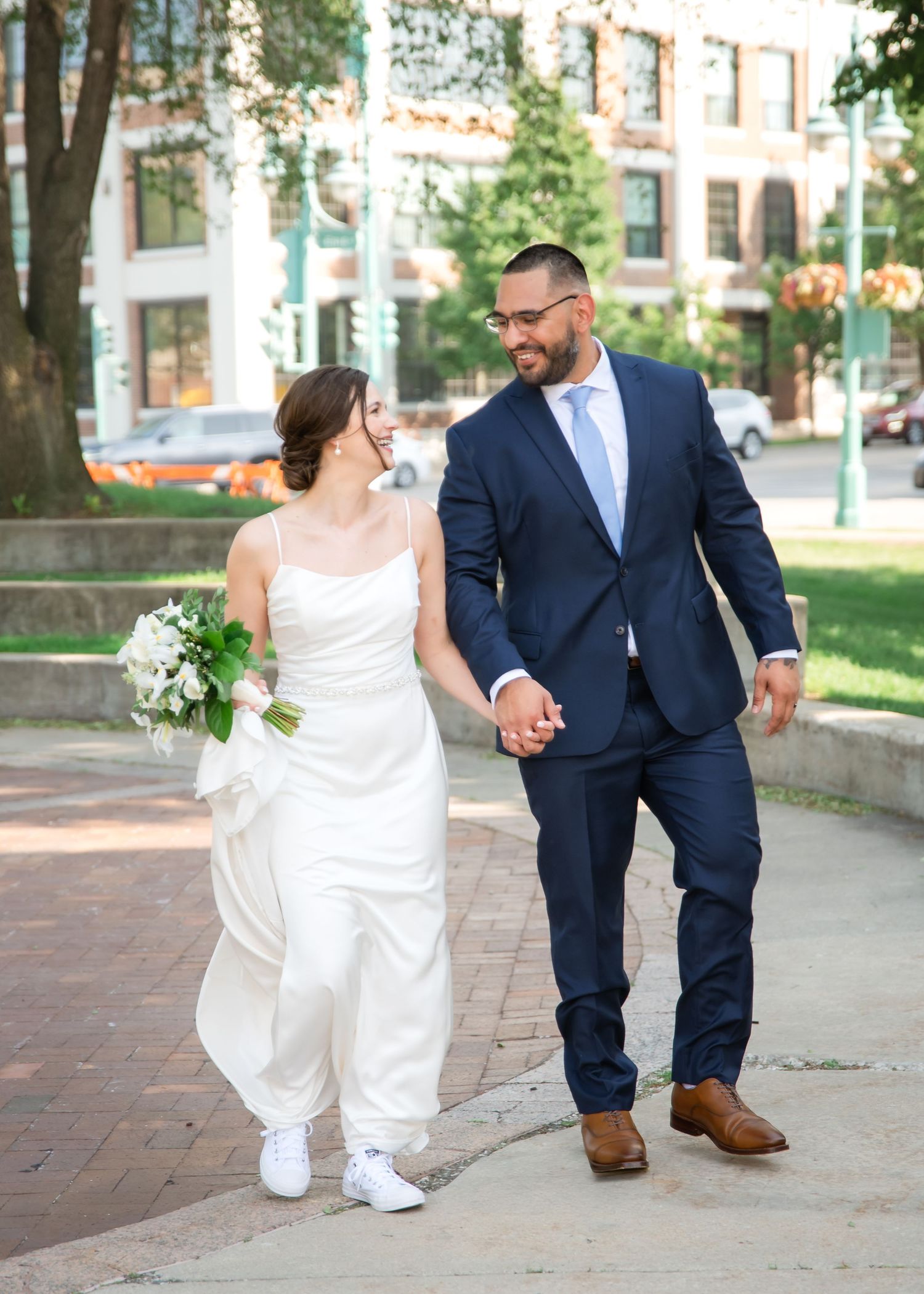 A couple in wedding attire walks together on a brick path, with the bride's dress flowing in the breeze.