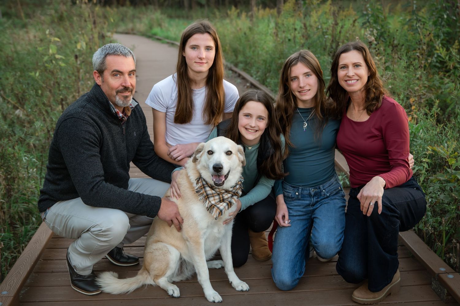 Family of five poses with golden retriever on wooden boardwalk path surrounded by lush greenery.