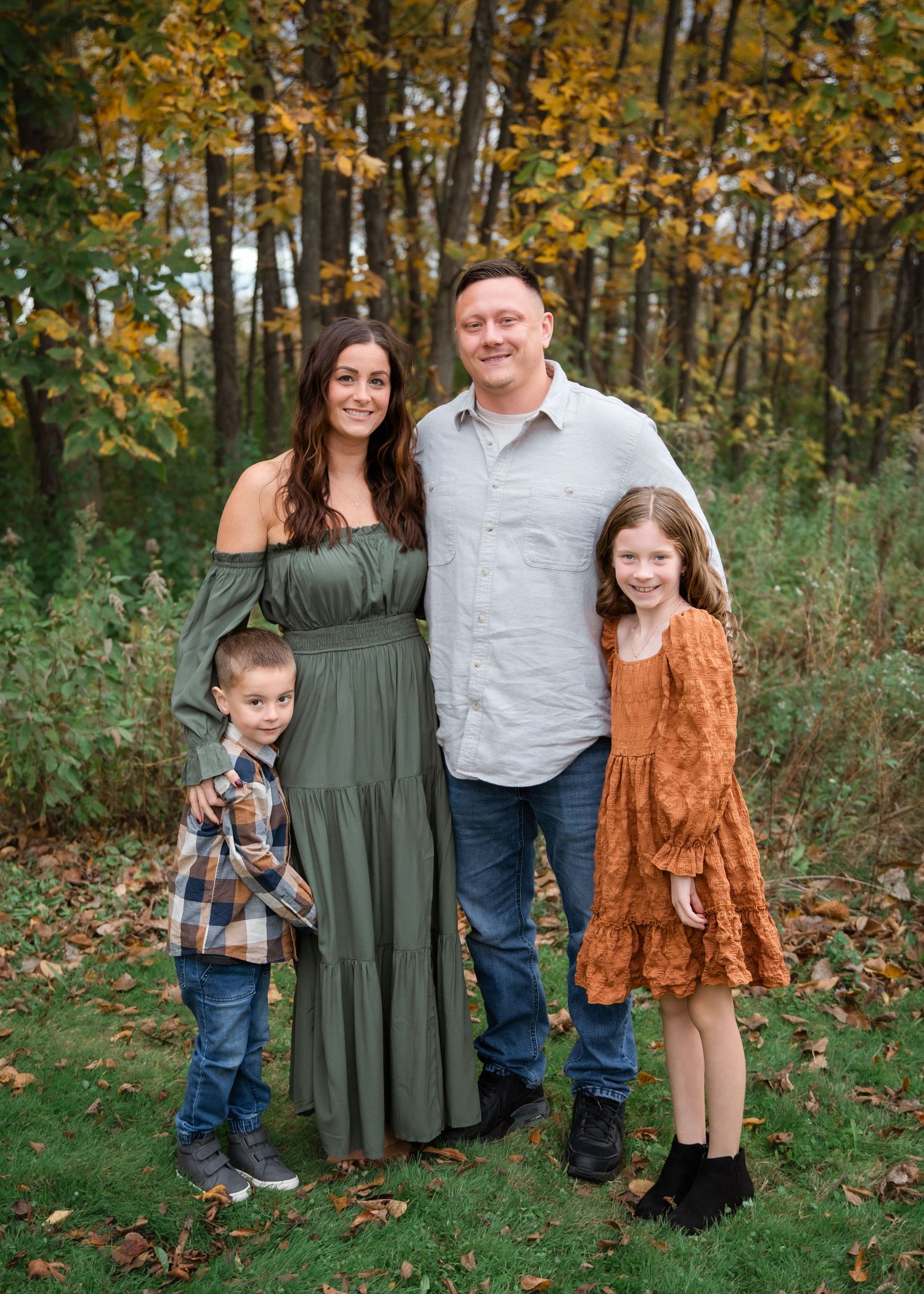 A family poses together outdoors in autumn, with colorful fall foliage in the background on a sunny day.