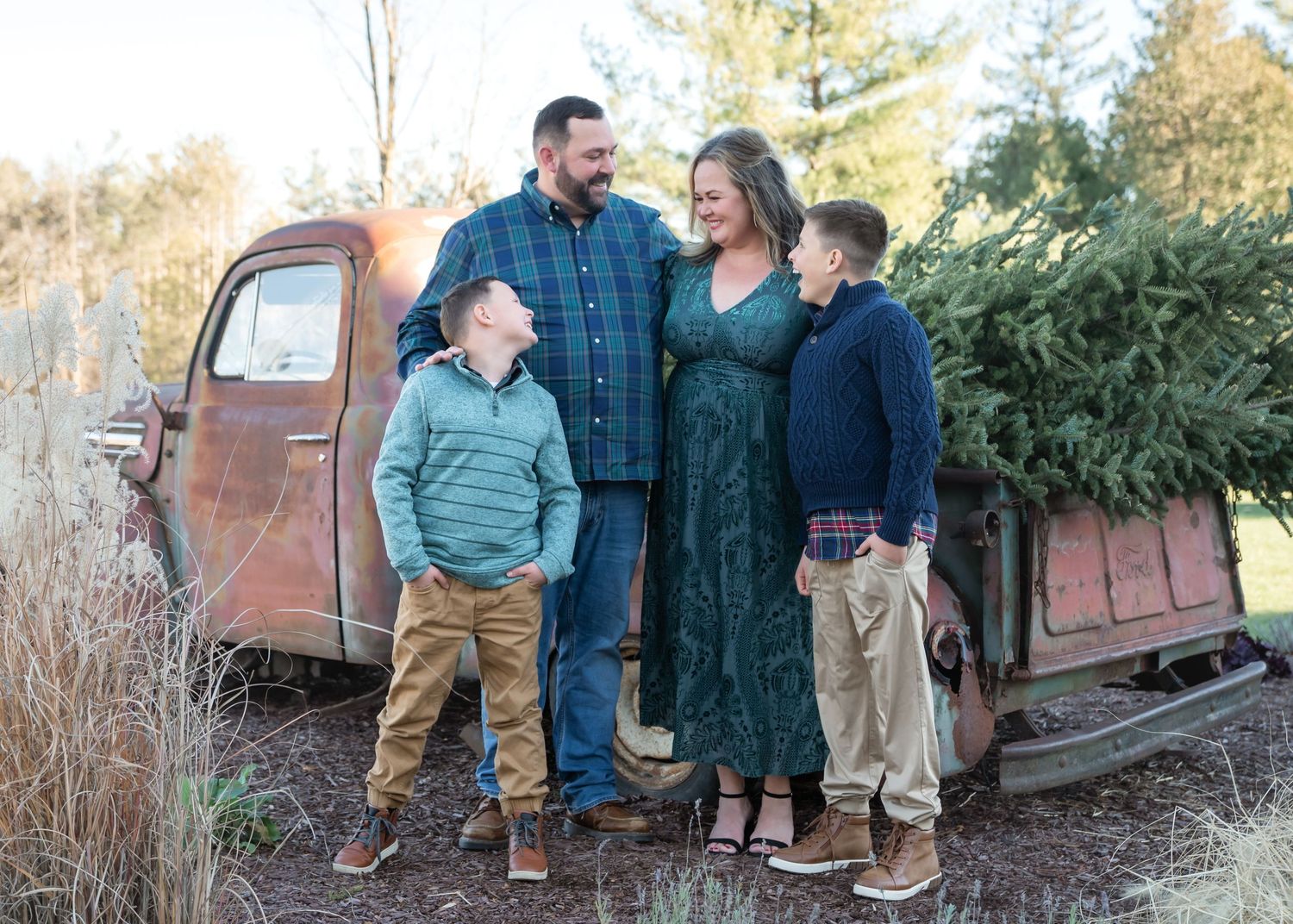 Family of four smiling together outdoors near a rustic vintage truck with a Christmas tree, dressed in holiday attire.