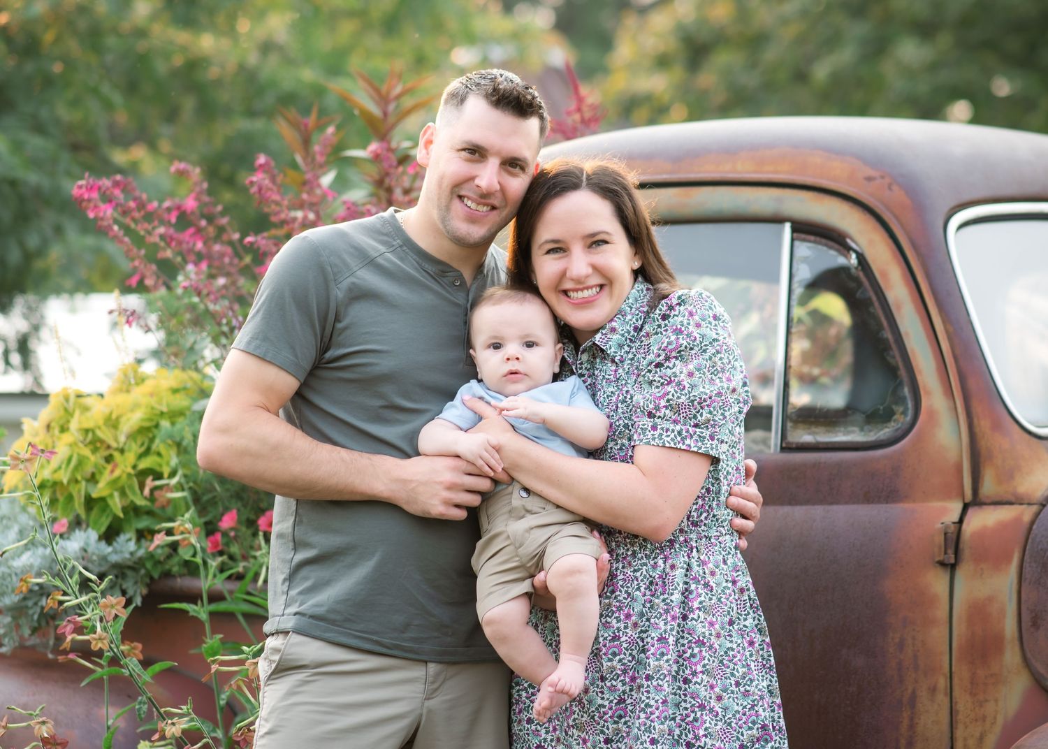 Happy young family with baby posing outdoors near a rustic vintage truck surrounded by lush garden greenery.