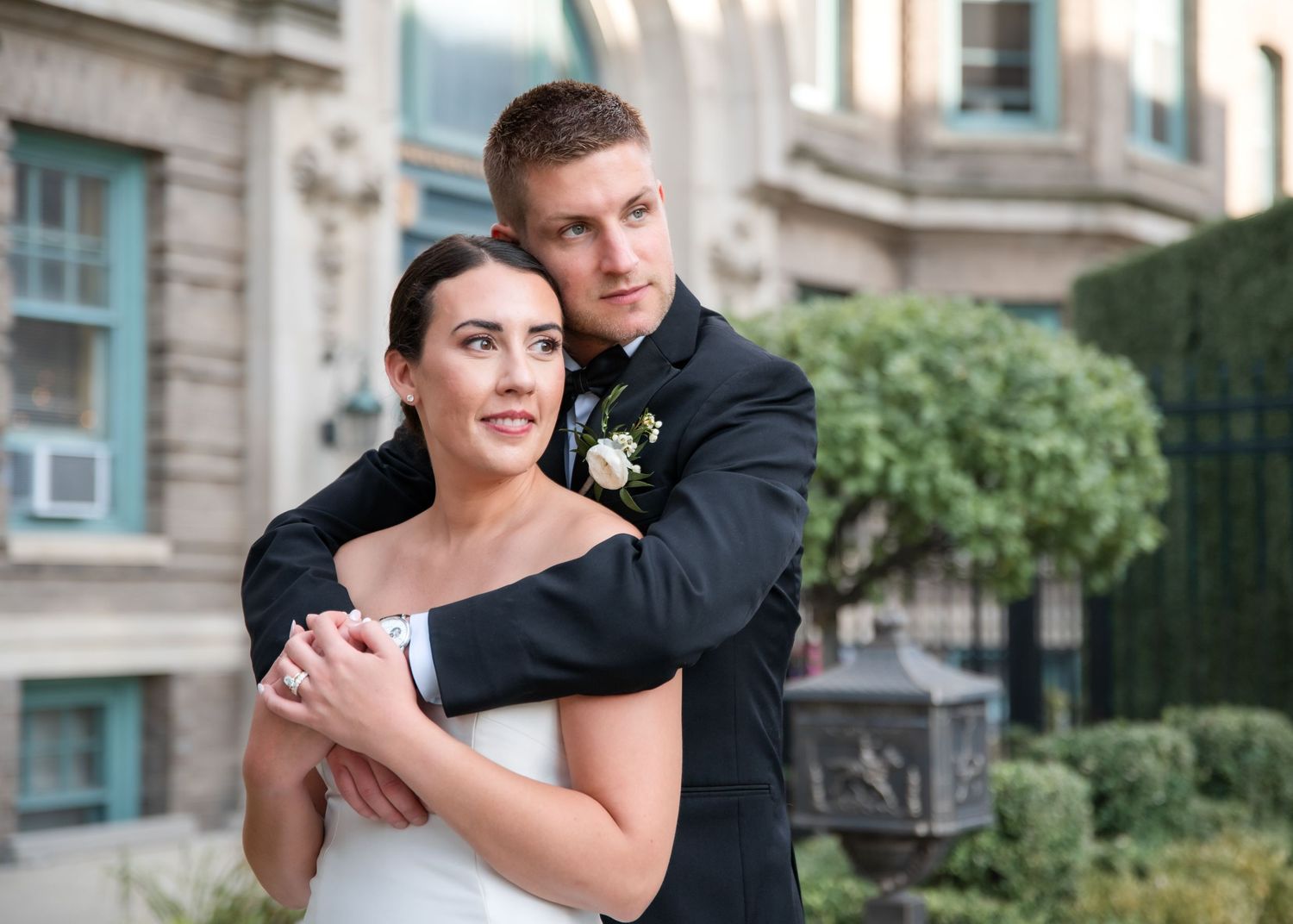 A couple in formal wedding attire embraces while posing for romantic outdoor portraits near a stone building.