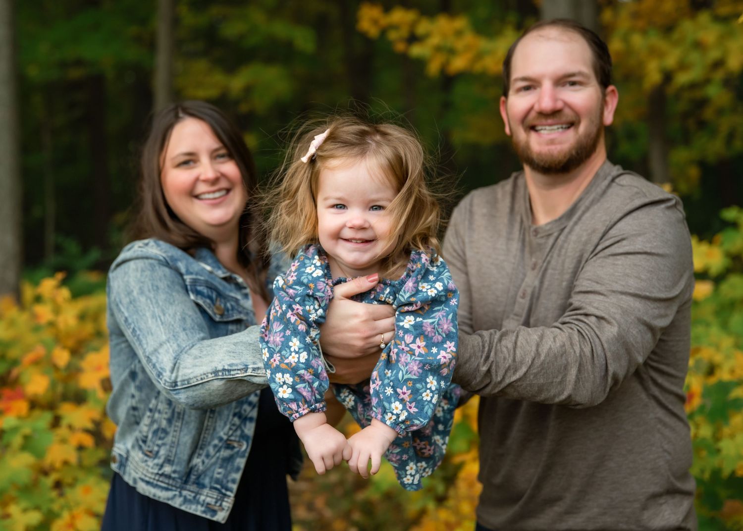 Happy family of three posing outdoors in autumn, parents holding smiling toddler girl among colorful fall foliage.