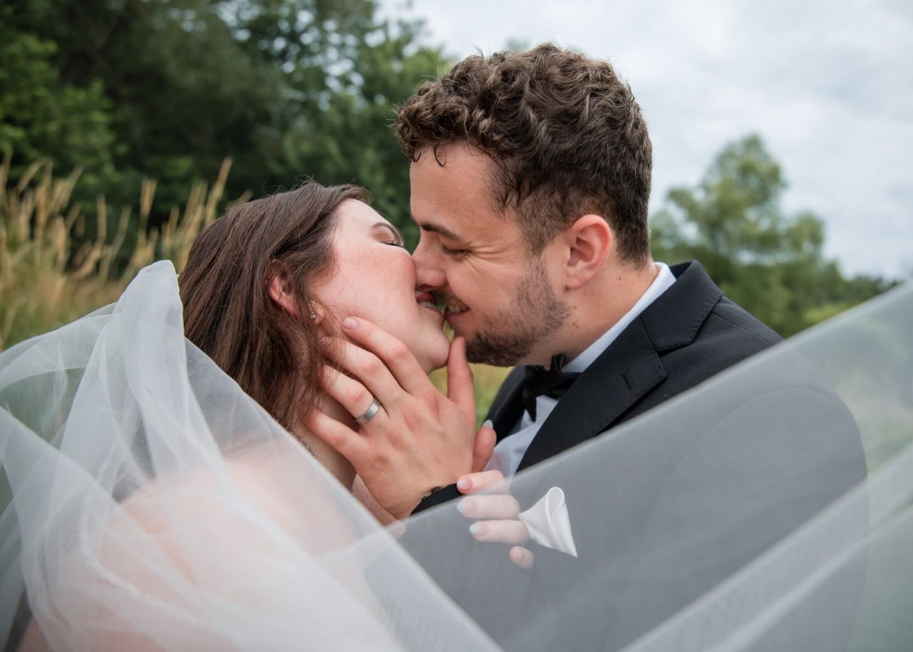 Tender kiss between newlyweds beneath flowing veil in natural setting.