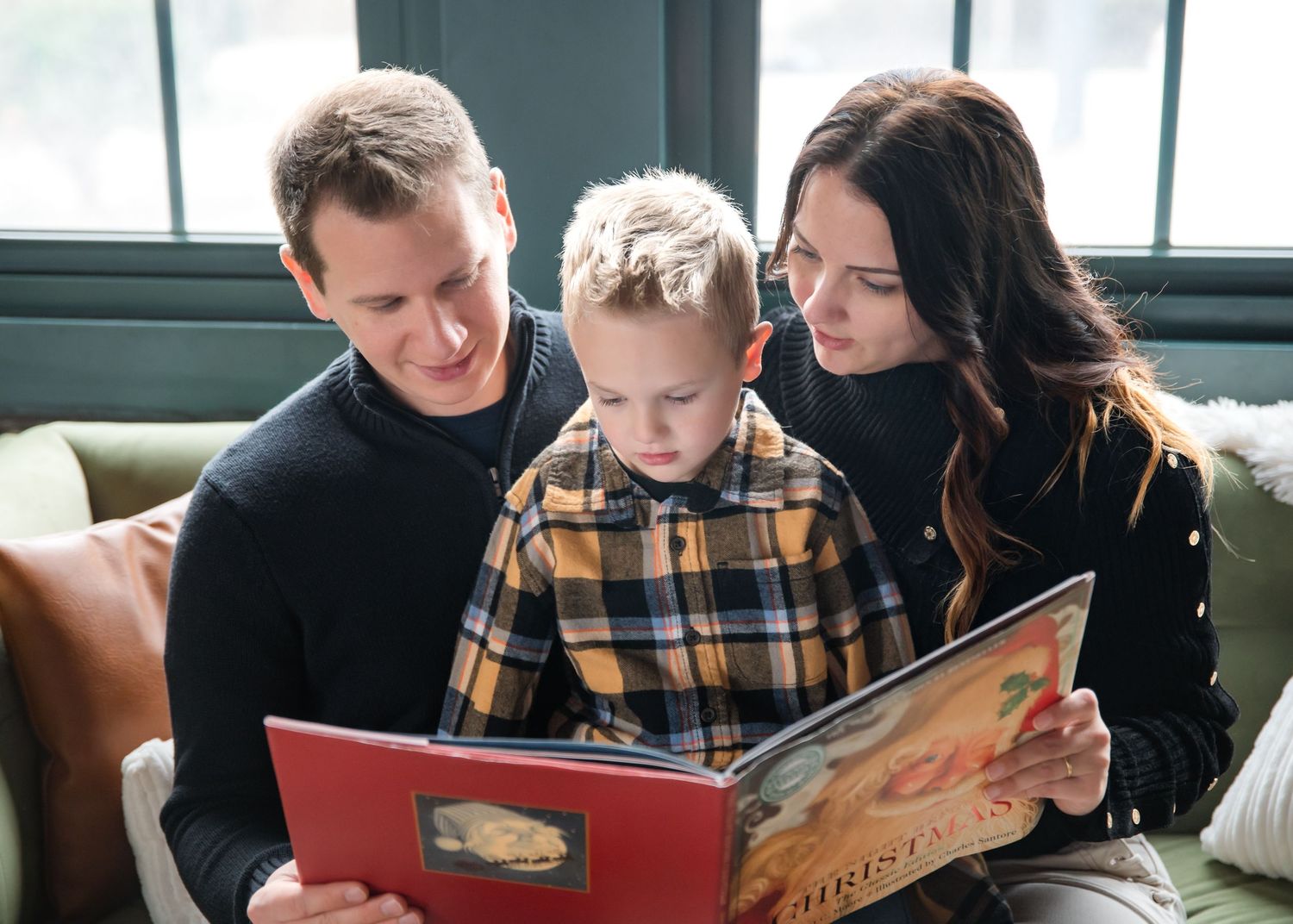 Family of three reading a Christmas book together on a couch, parents and young boy enjoying holiday storytime.