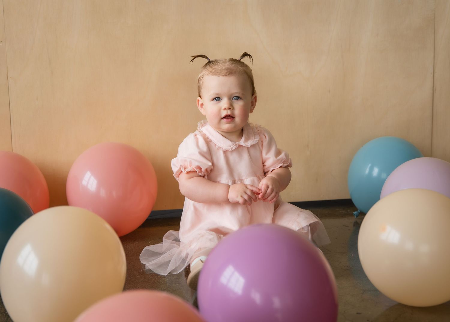 Baby girl in pink dress sits surrounded by colorful balloons, celebrating her first birthday party.