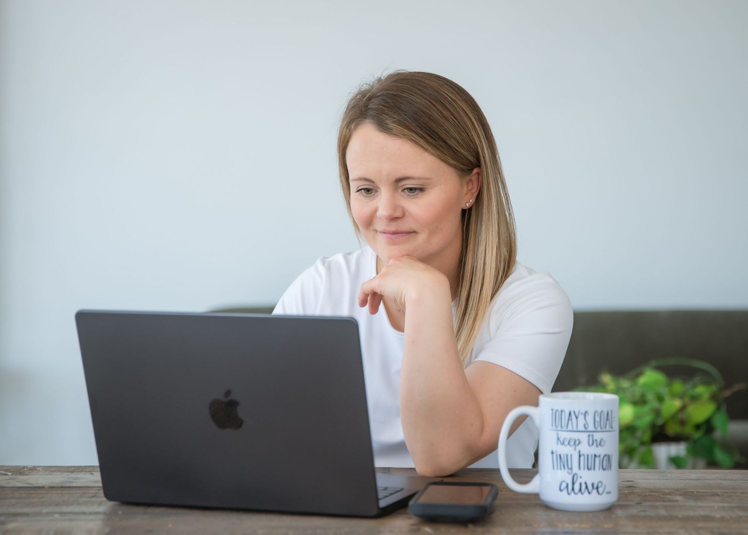 Woman smiling while working on a MacBook laptop at a wooden desk with a motivational mug and plant nearby.