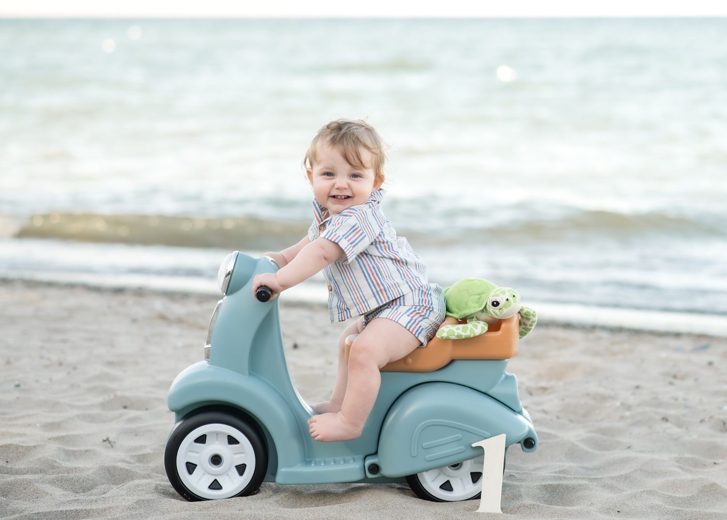 Toddler riding blue toy scooter on beach with stuffed animal.