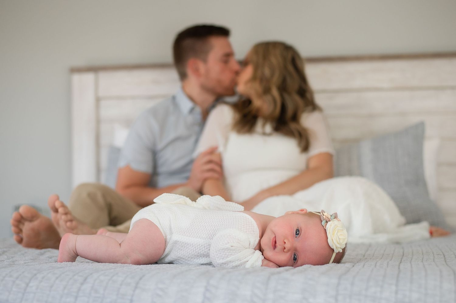 Newborn baby sleeps peacefully on bed while parents share tender moment in background.