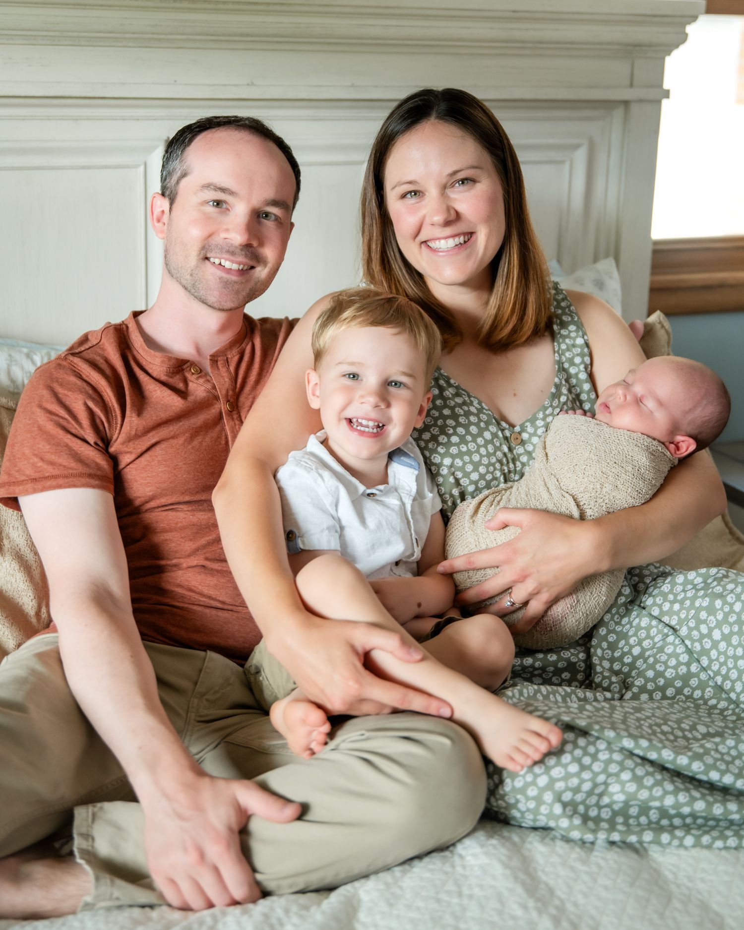 Family portrait on bed with parents, toddler and newborn in casual attire.