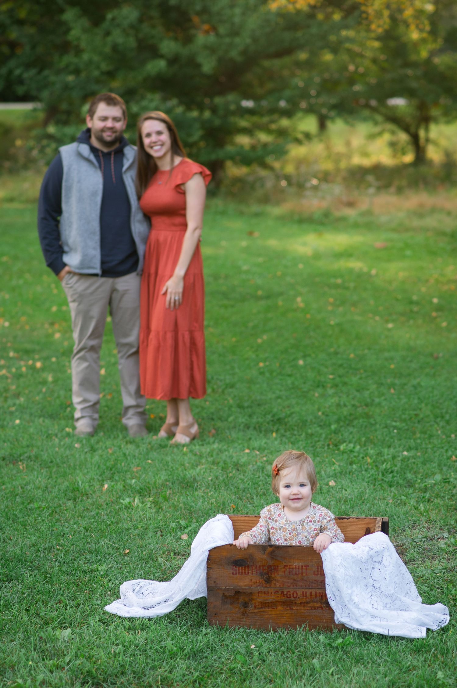 Family portrait in a garden with baby sitting in wooden crate while parents stand together on green grass.