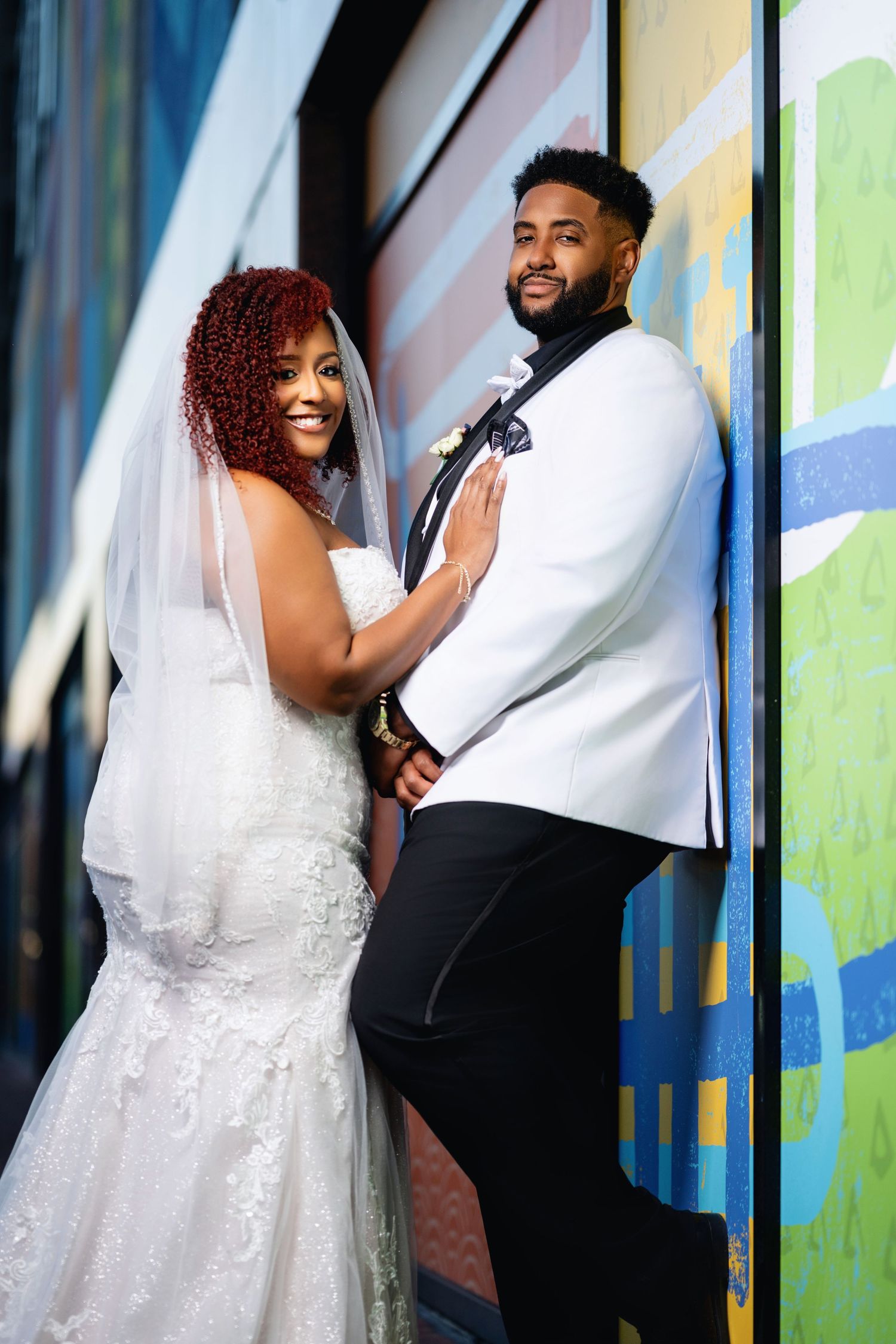 A couple in wedding attire poses against a colorful mural wall, the bride in a lace gown and veil, groom in white jacket.