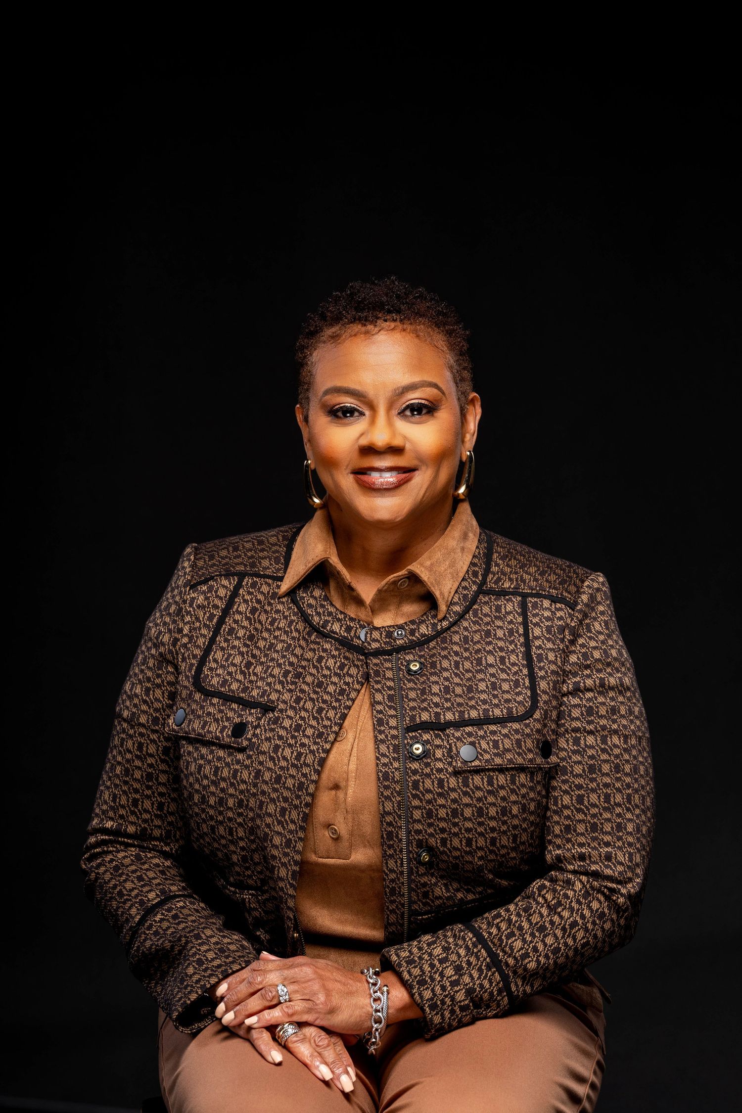 Professional woman in brown patterned blazer smiling confidently against dark background in formal portrait.