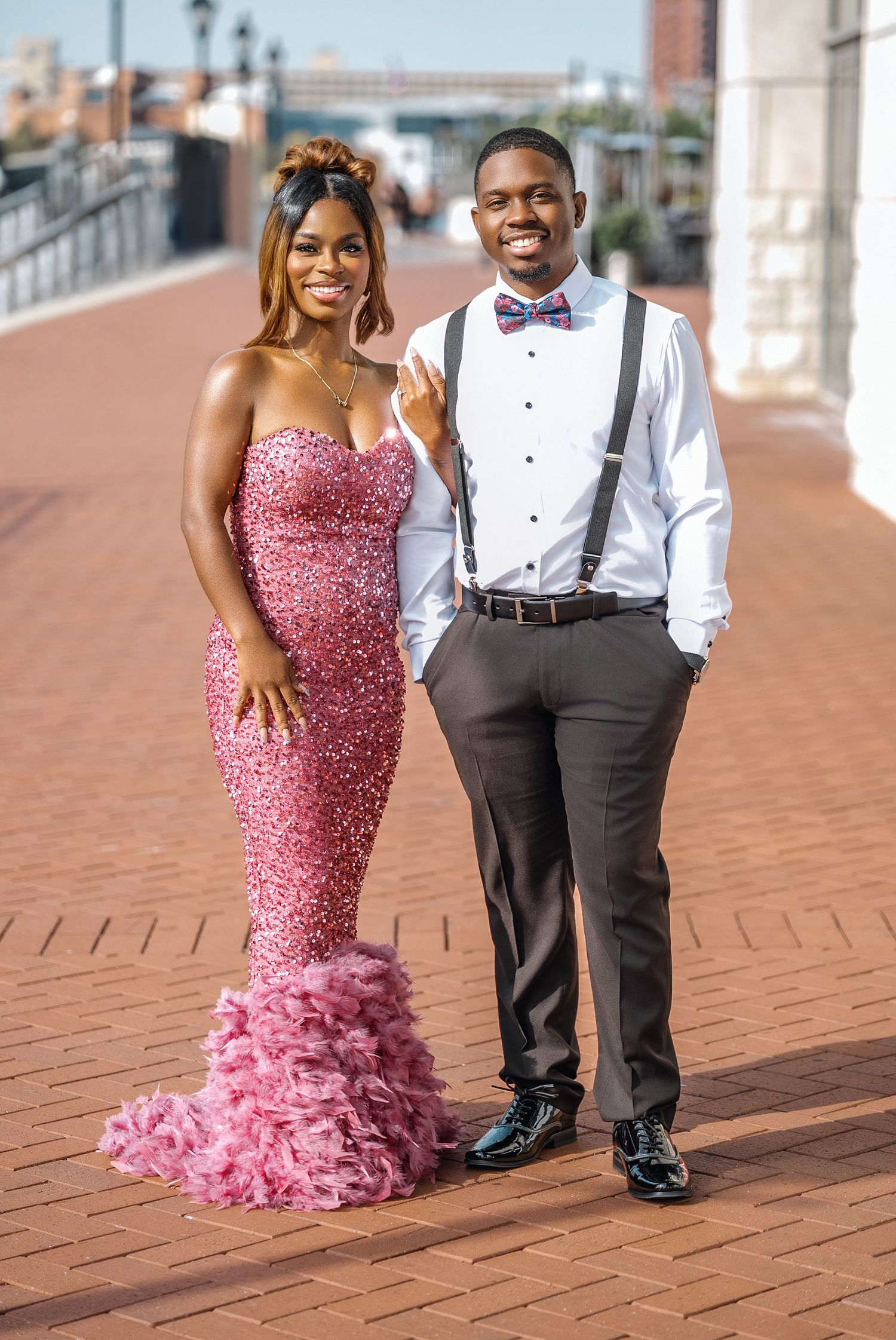 A couple poses together on brick pavement, with one wearing a sparkly pink gown and the other in formal attire.