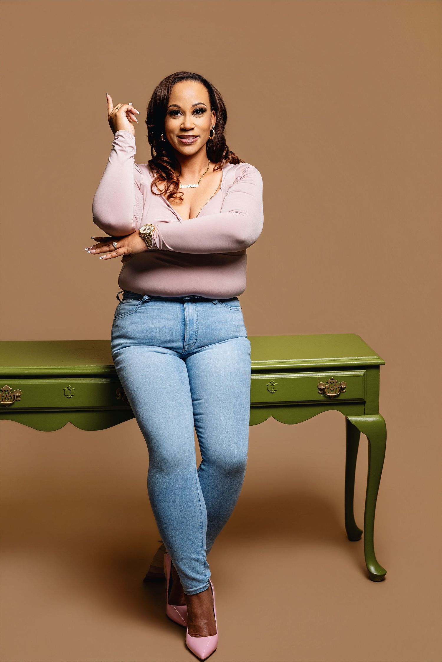 Woman in pink top and jeans leaning on green desk, posing confidently against a warm brown background.
