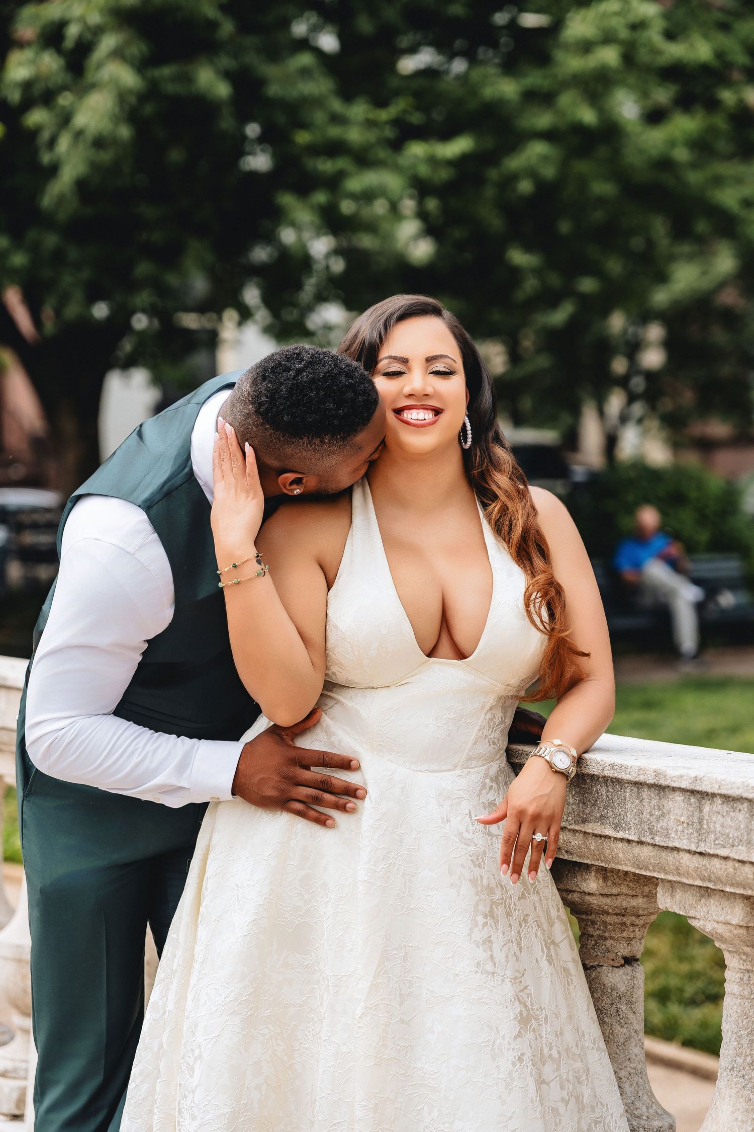 Groom kissing bride on cheek as she smiles joyfully, wearing a deep V-neck wedding gown outdoors.