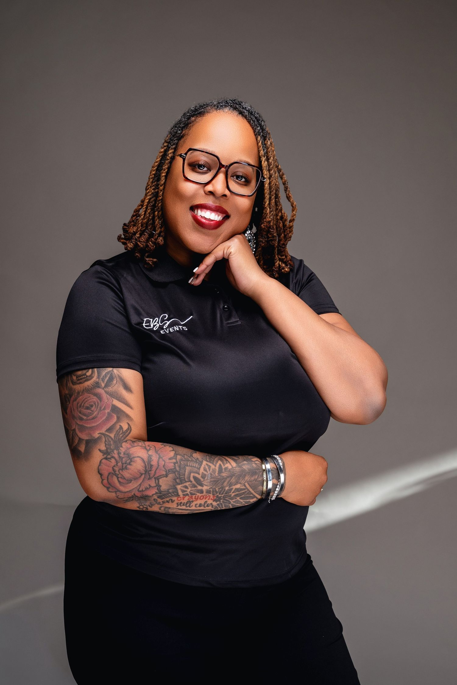 Woman in black chef coat with floral arm tattoos, glasses, and braids poses confidently in professional studio portrait.
