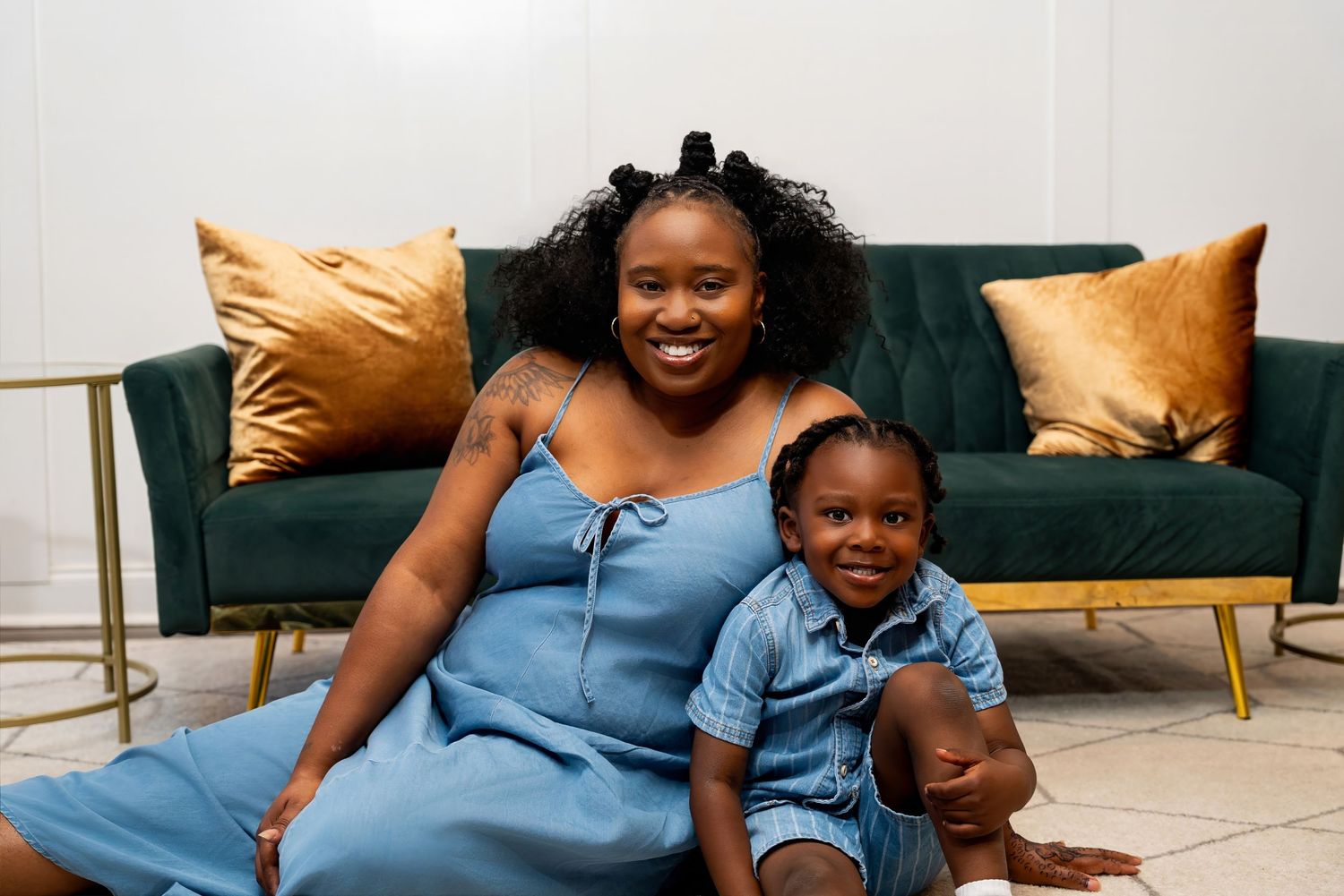 A smiling woman and young child in matching blue outfits sit together in front of a green velvet sofa.