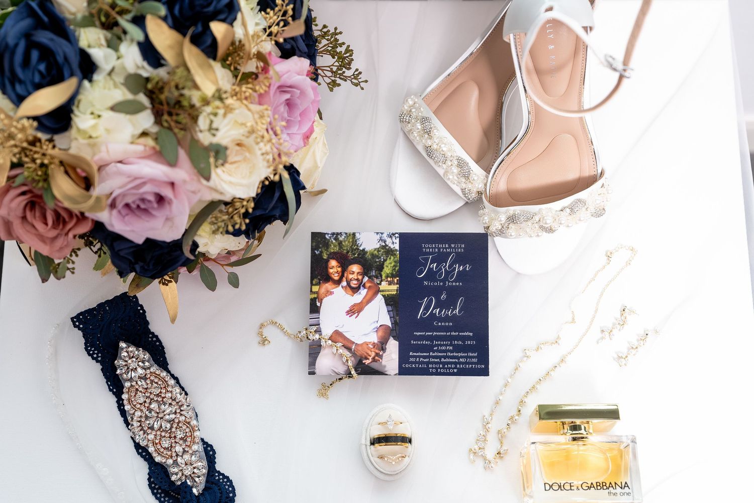 Wedding details flatlay with navy blue invitation, rose bouquet, crystal heels, garter and perfume on marble surface.