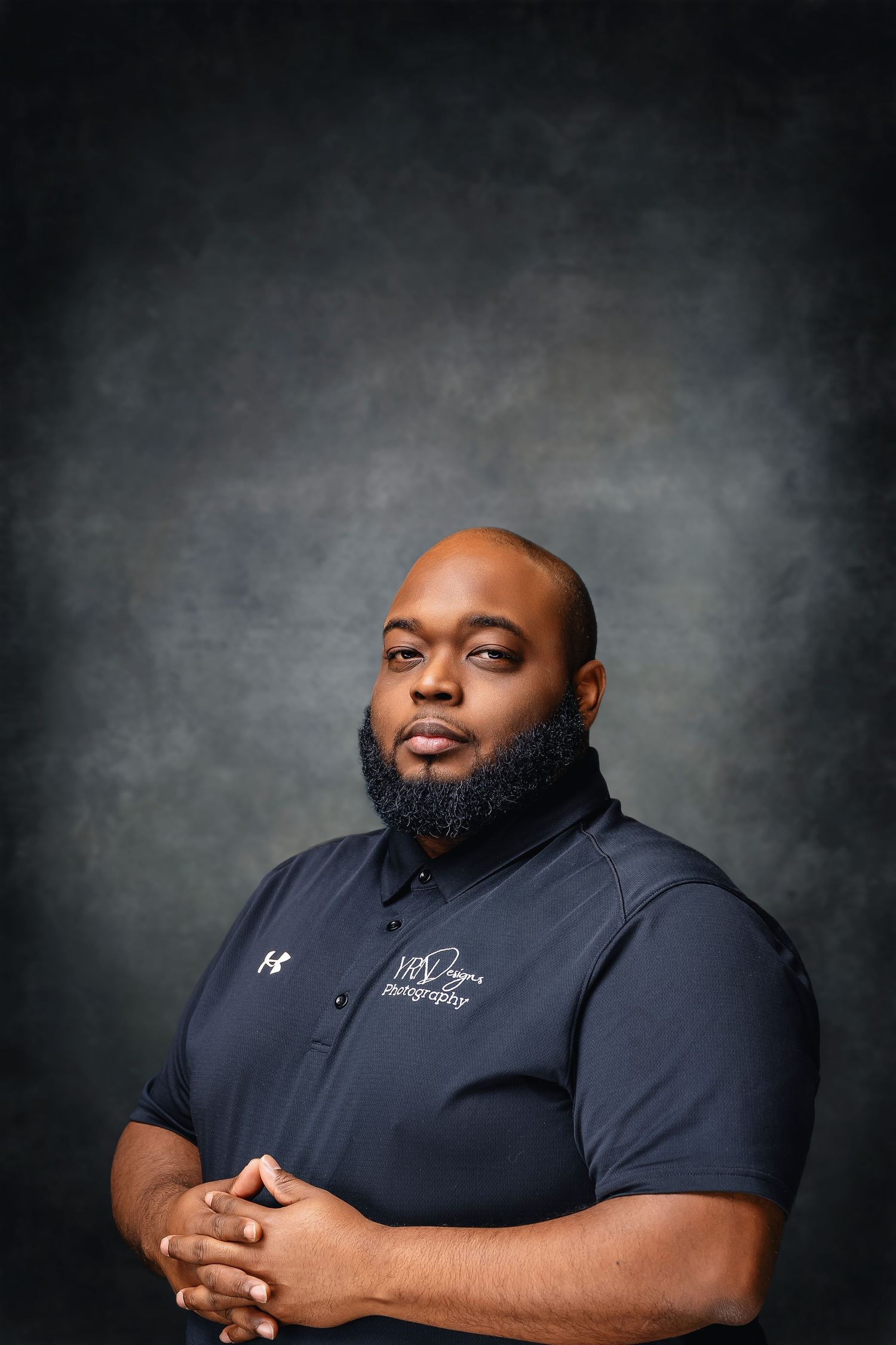 Professional studio portrait of person in navy Under Armour polo against dark gray background with dramatic lighting.
