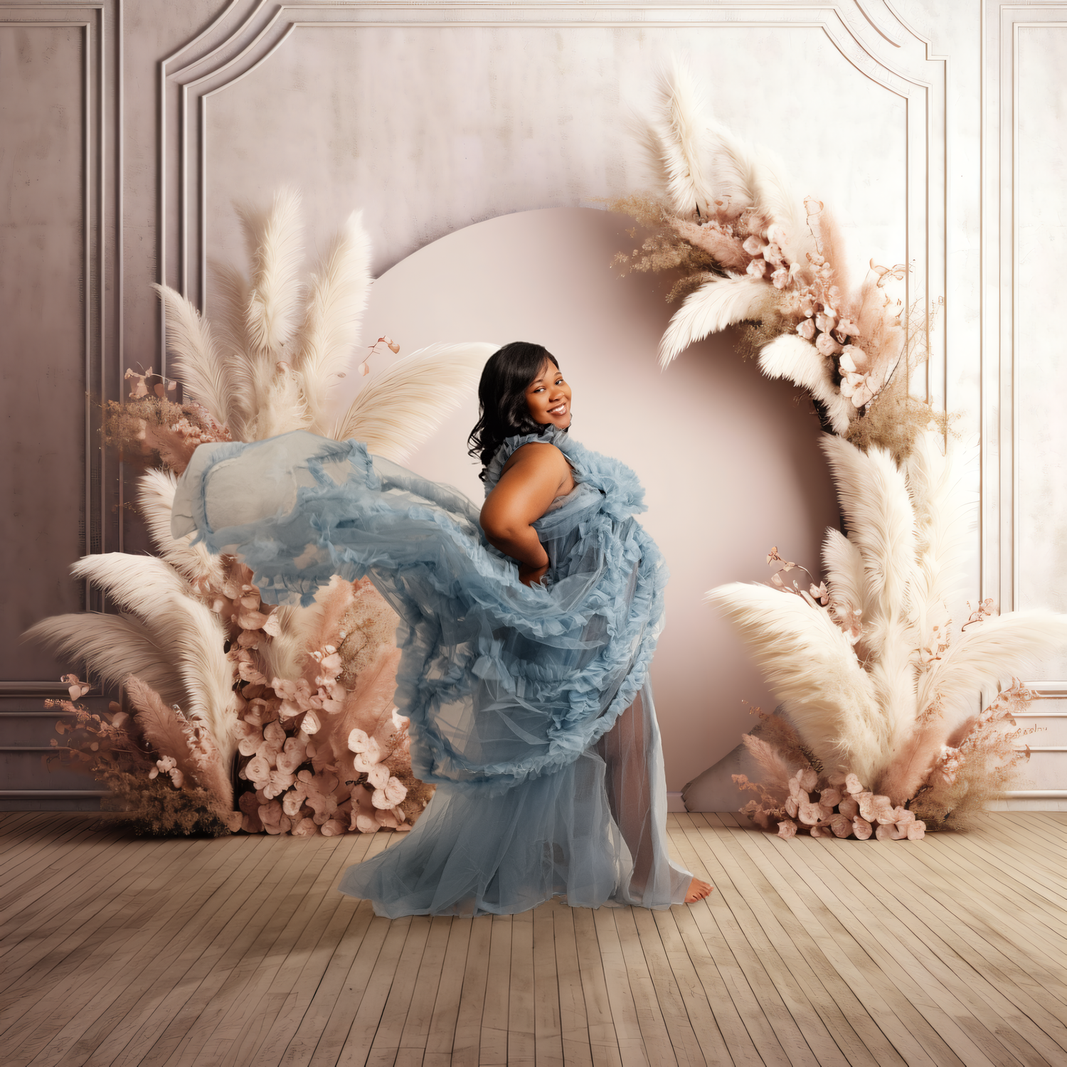 Pregnant woman in blue tulle gown poses in floral arch with pampas grass for elegant maternity photo.