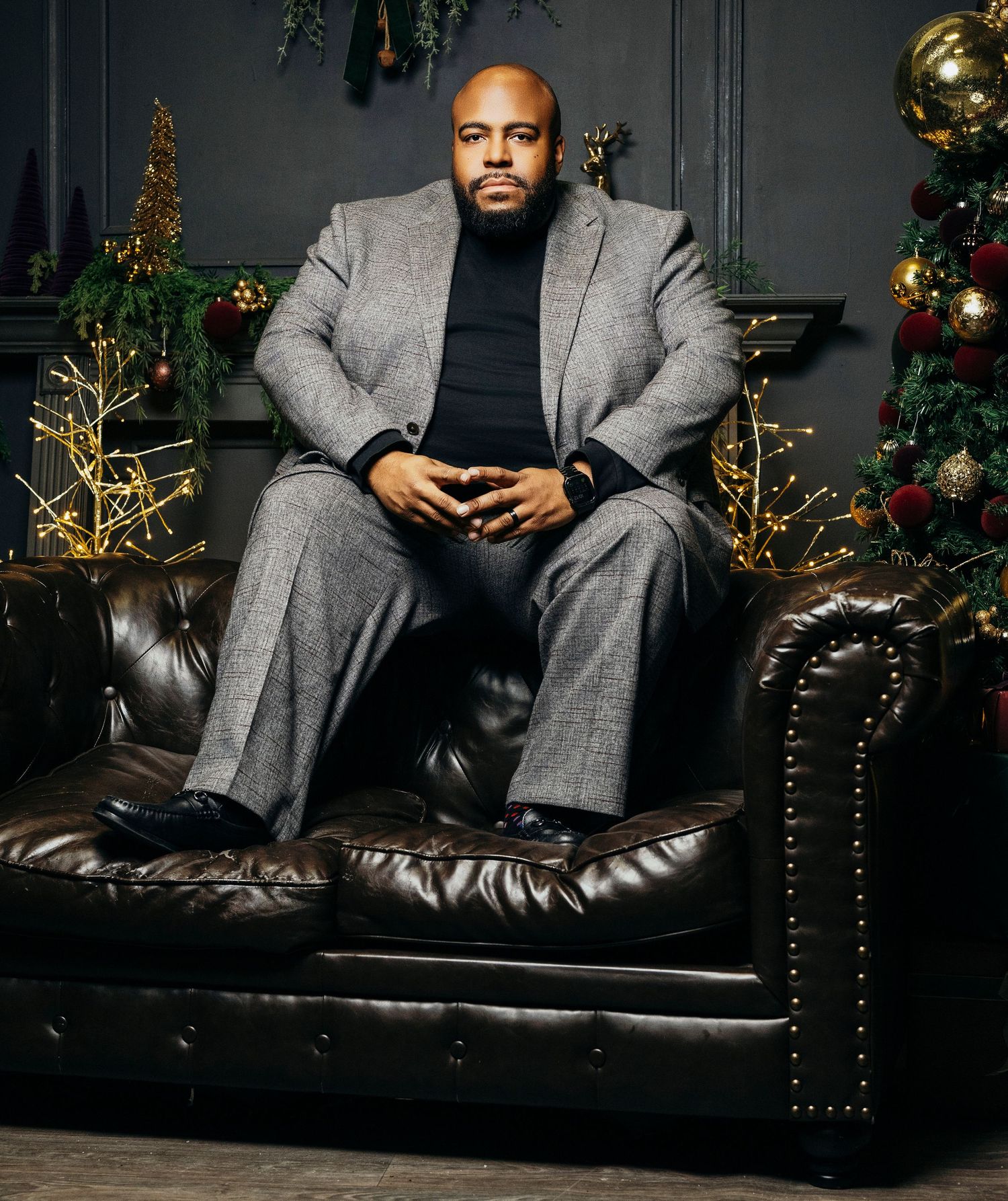 A person in a grey suit and black shirt lounges on a dark leather couch with Christmas decorations in the background.