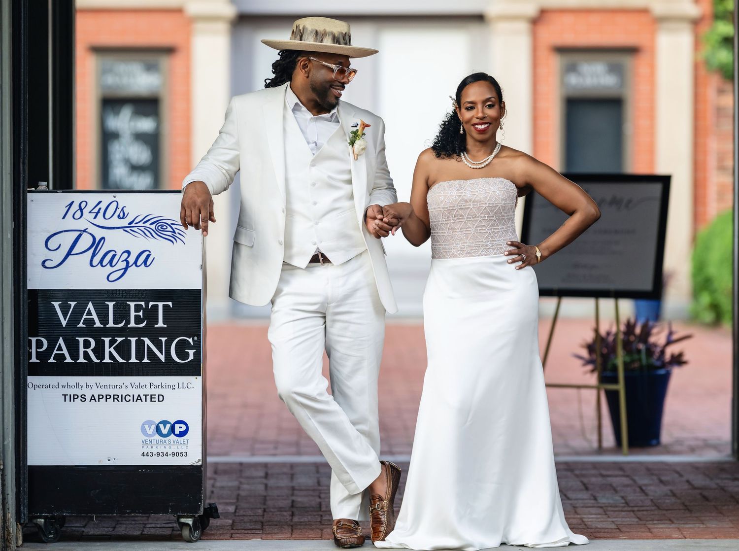 A couple in elegant white outfits dances together on a brick walkway near valet parking signs.
