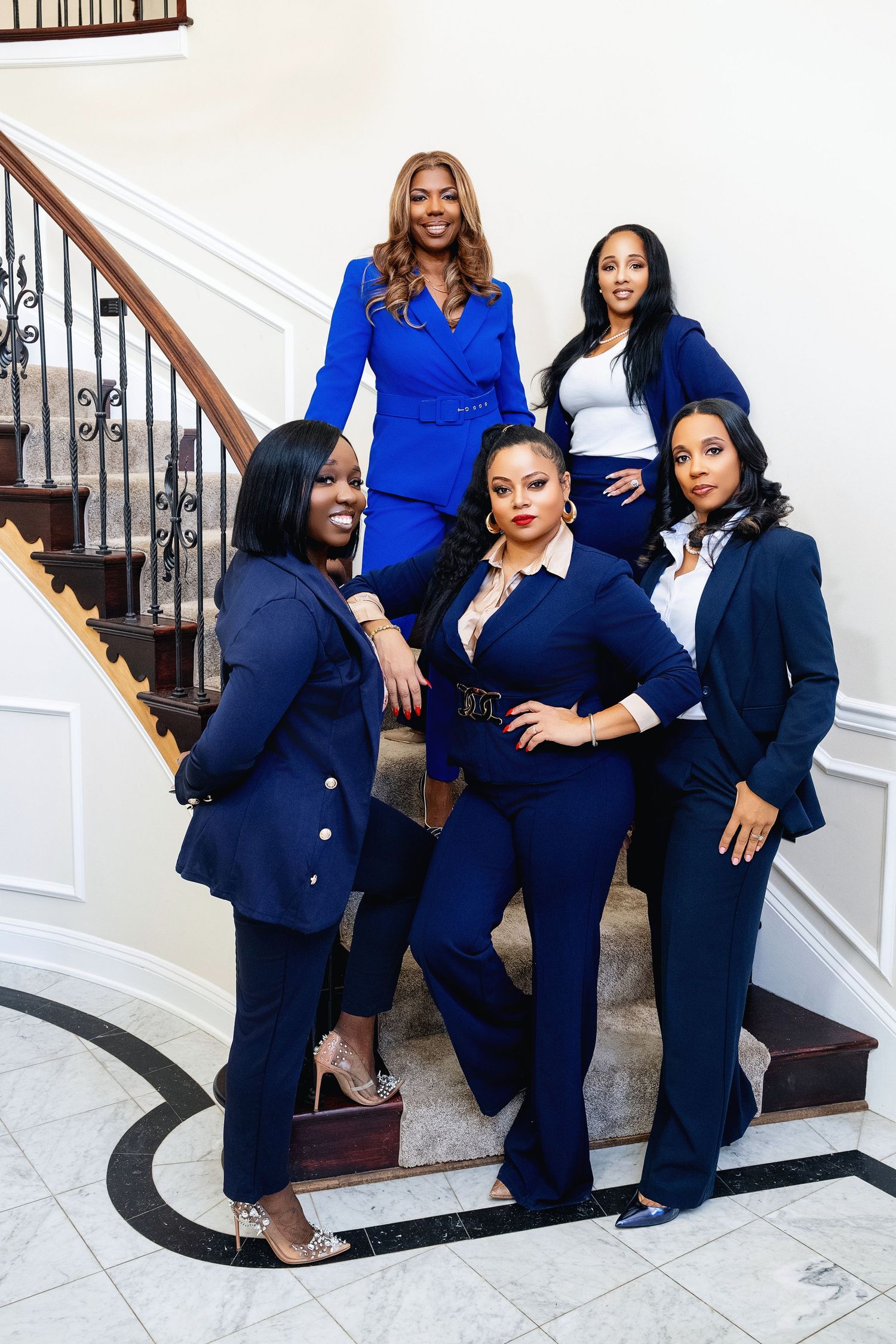 Five professional women in coordinating blue suits pose confidently on an elegant staircase in a formal setting.