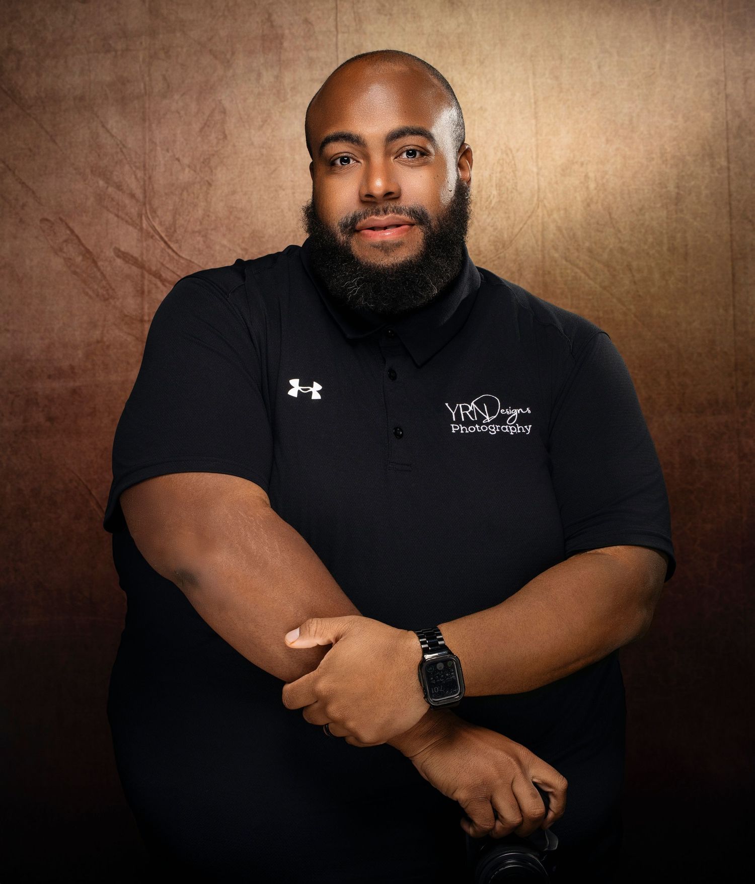 Professional portrait against textured brown backdrop showing person in black Under Armour shirt with crossed arms.