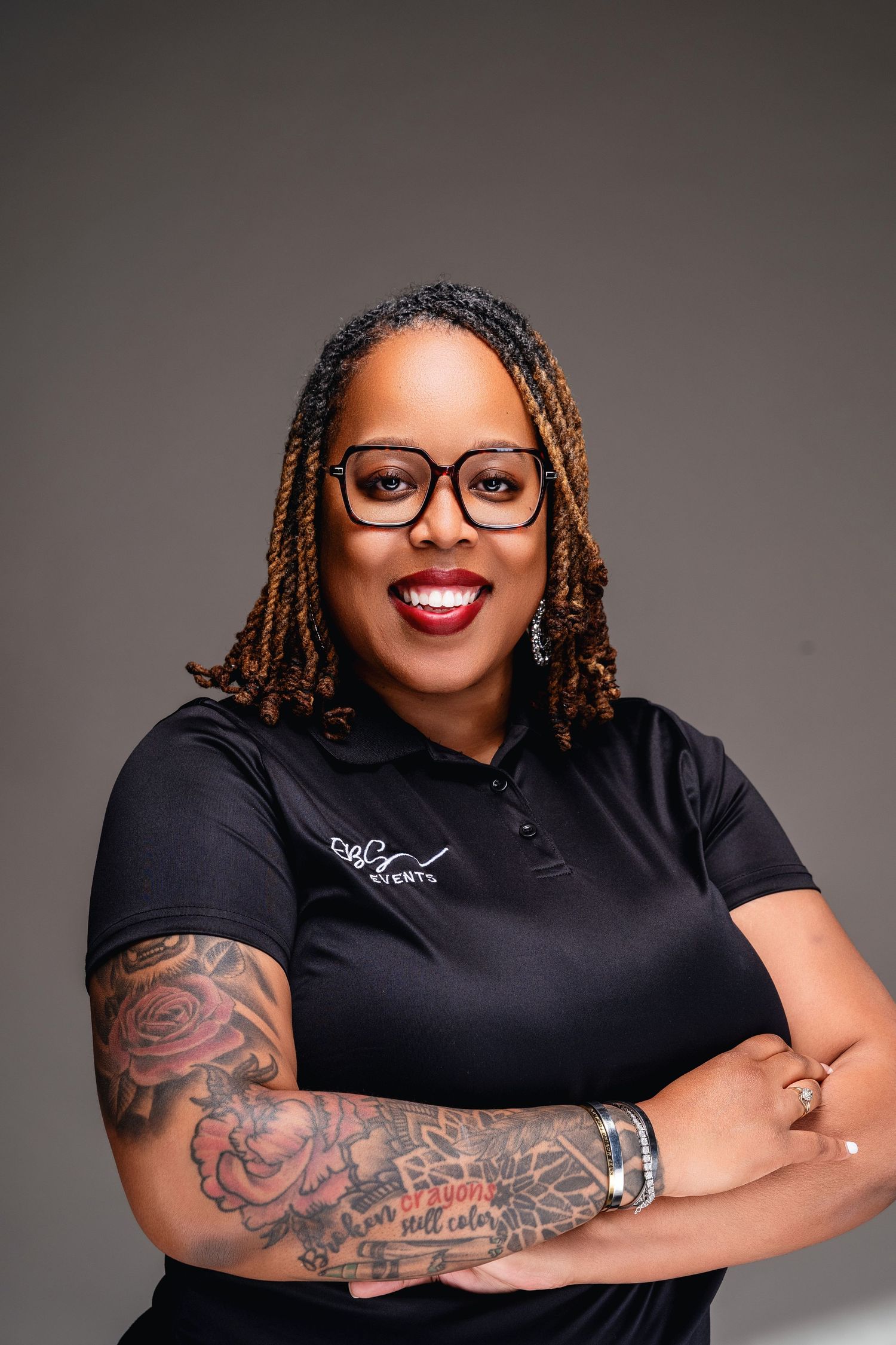 Professional woman with braids, glasses, and tattoos poses confidently with arms crossed wearing a black polo shirt.