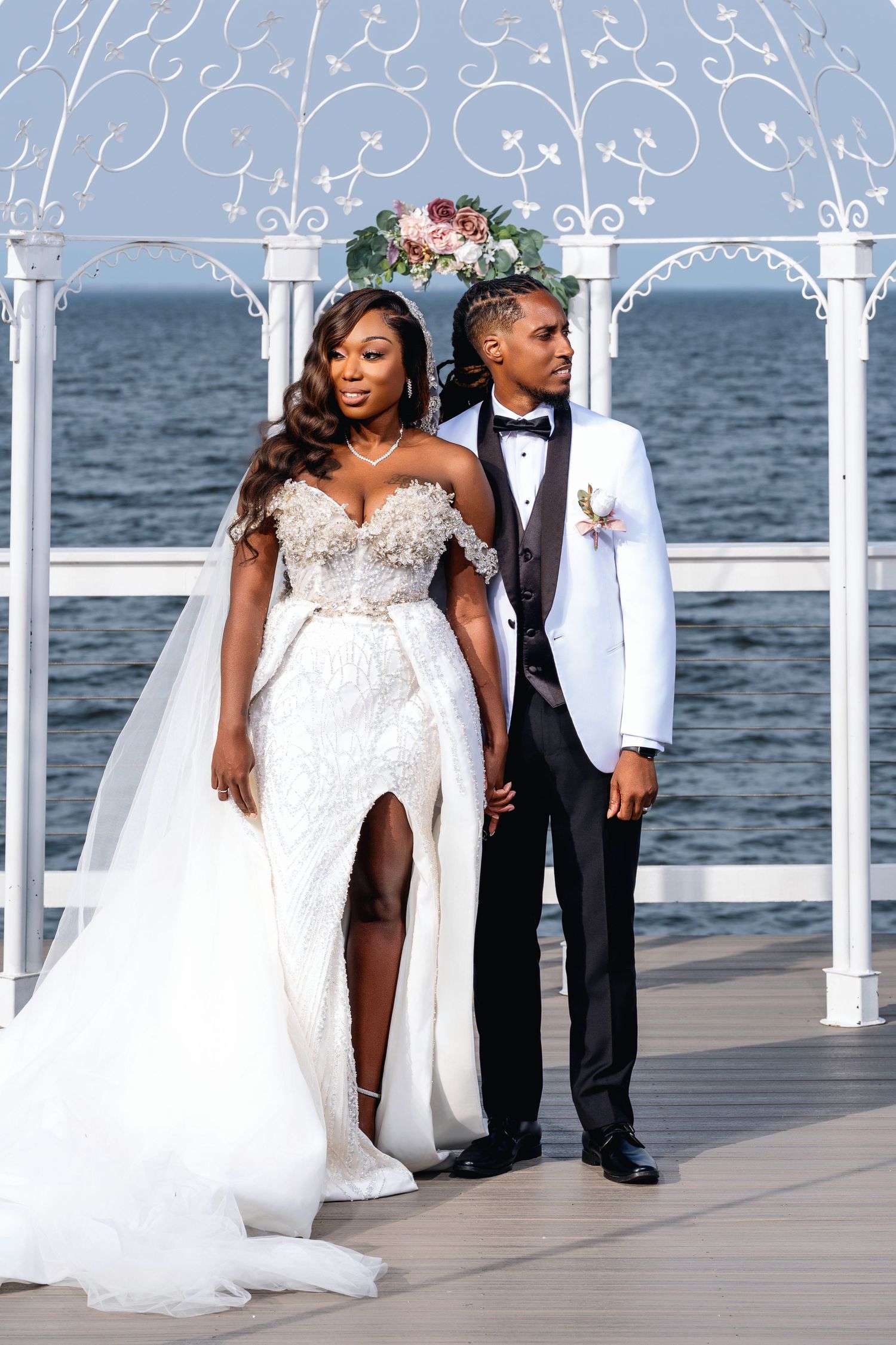 A couple poses at a waterfront wedding ceremony under an ornate white arch with floral decorations.