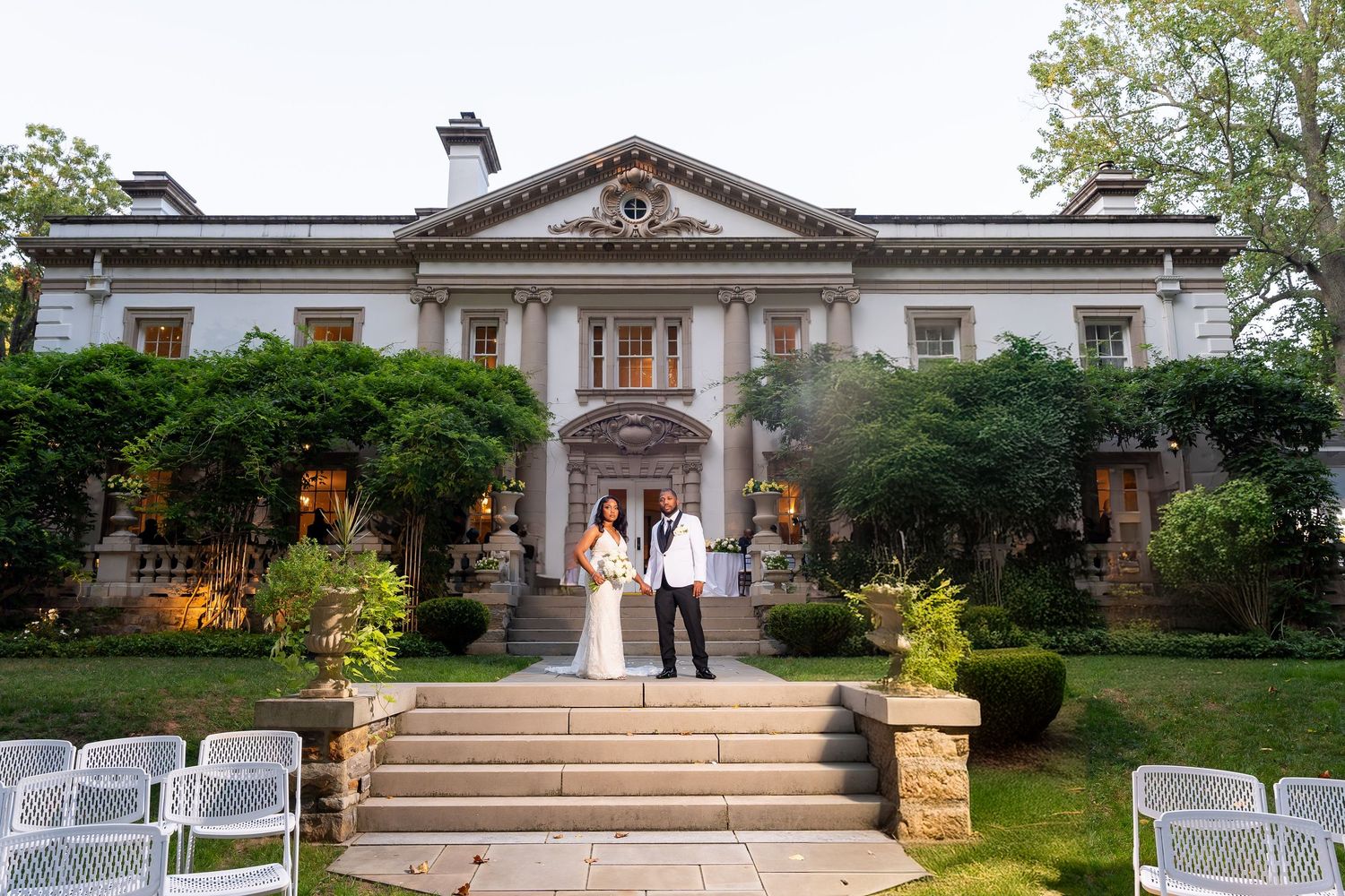 A grand mansion wedding venue with stone steps, classic architectural details, manicured gardens and white folding chairs.