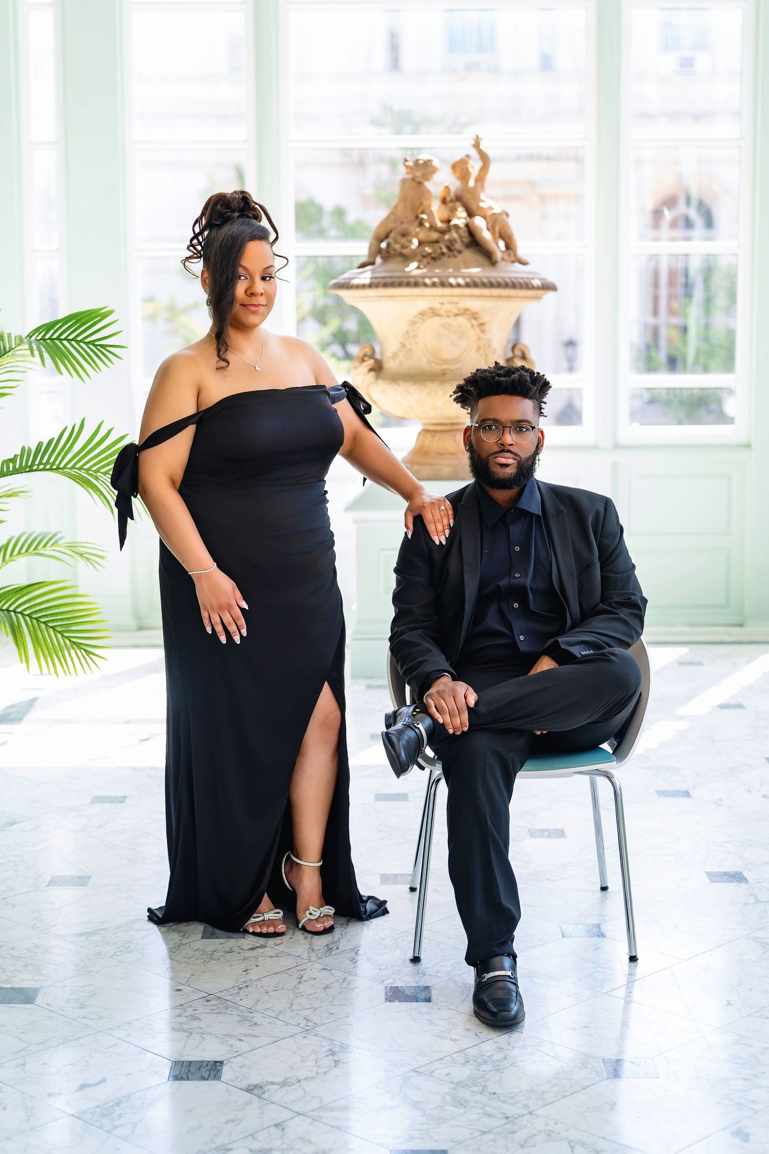 A couple in elegant black attire poses together in a bright sunlit room with tropical plants and modern decor.