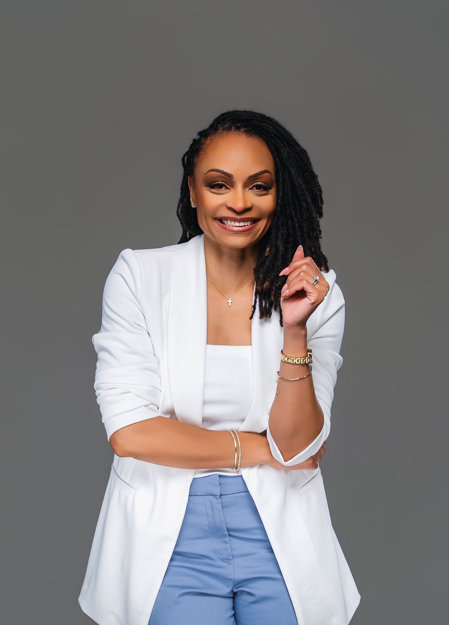 Professional wearing a crisp white blazer and blue pants poses confidently against a gray studio backdrop.